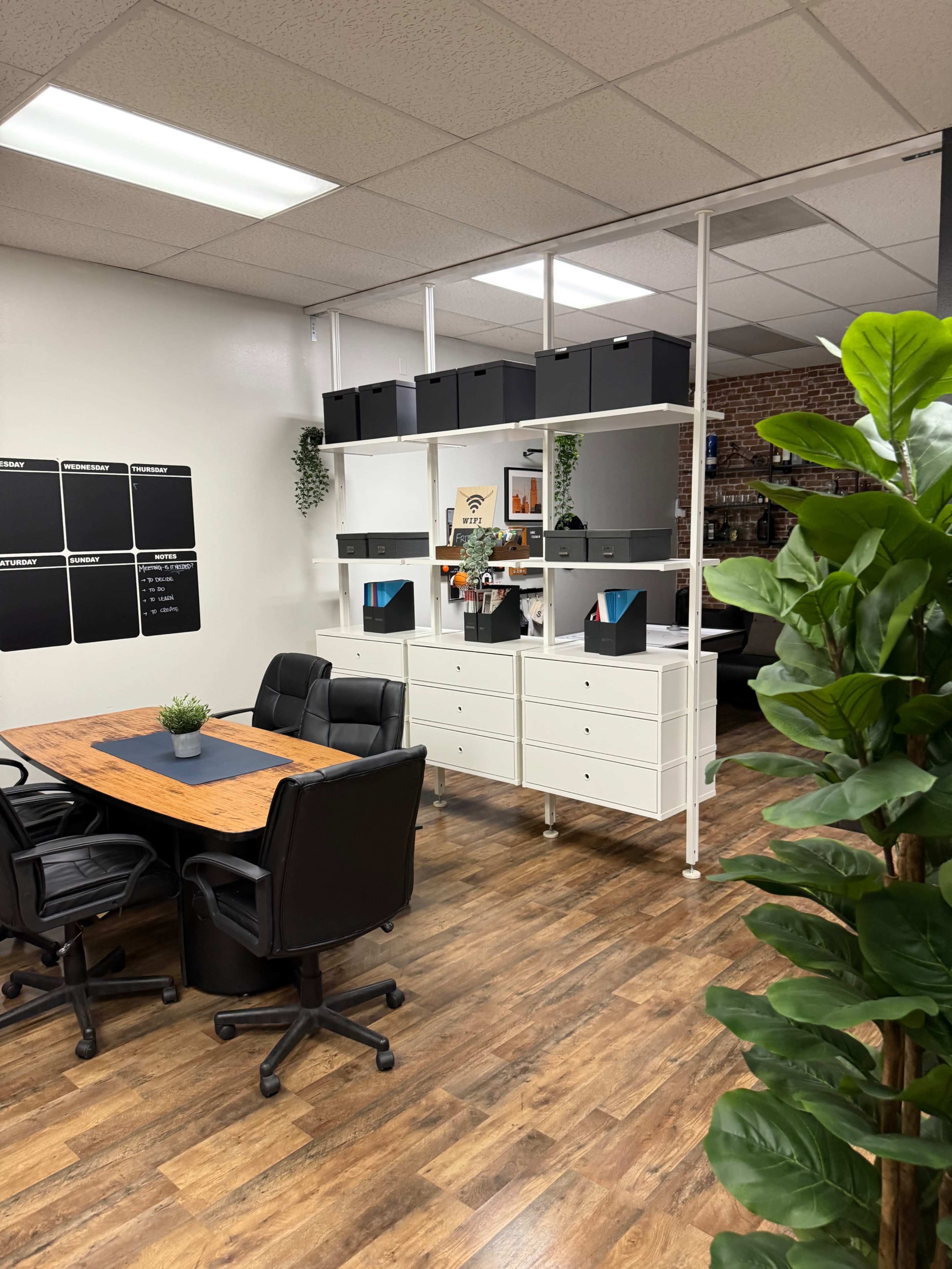 The image shows a modern office space divided by a shelving unit, featuring a table with black chairs and storage boxes on the shelves.