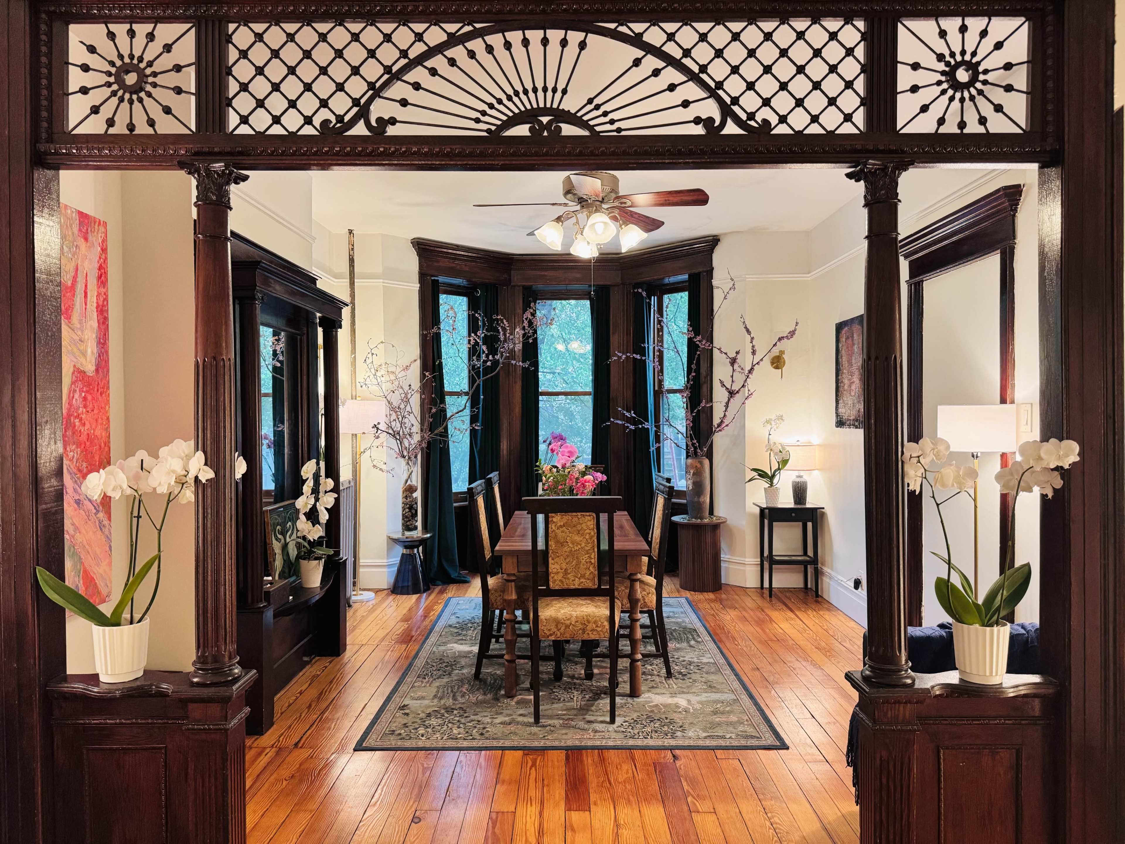 The image shows a dining room with a wooden table and chairs, framed by an ornate wooden archway, and large windows allowing natural light to fill the space.