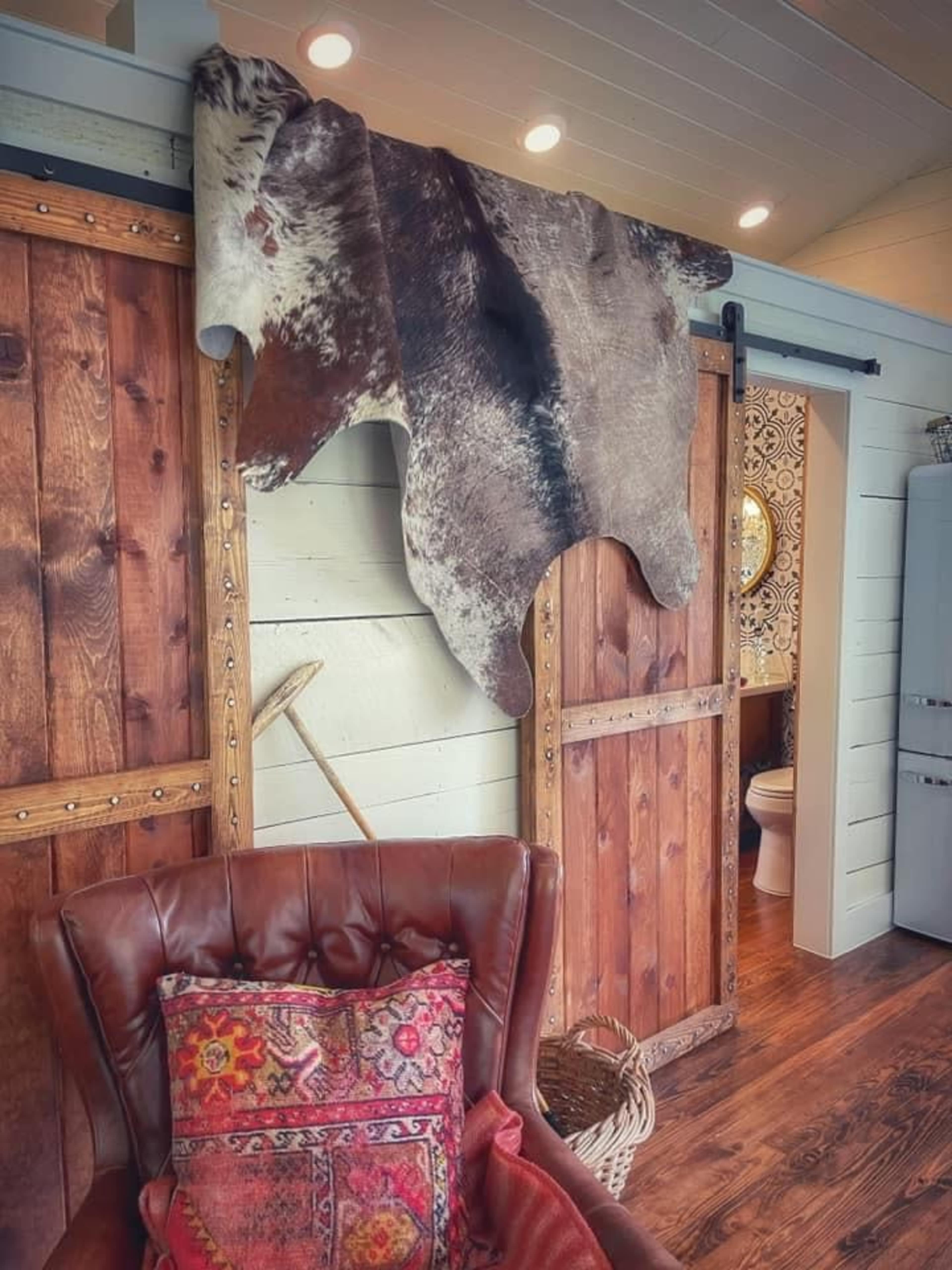 A cowhide rug hangs on a wooden wall above a brown leather armchair, with a decorative pillow and visible wooden doors leading to a bathroom.