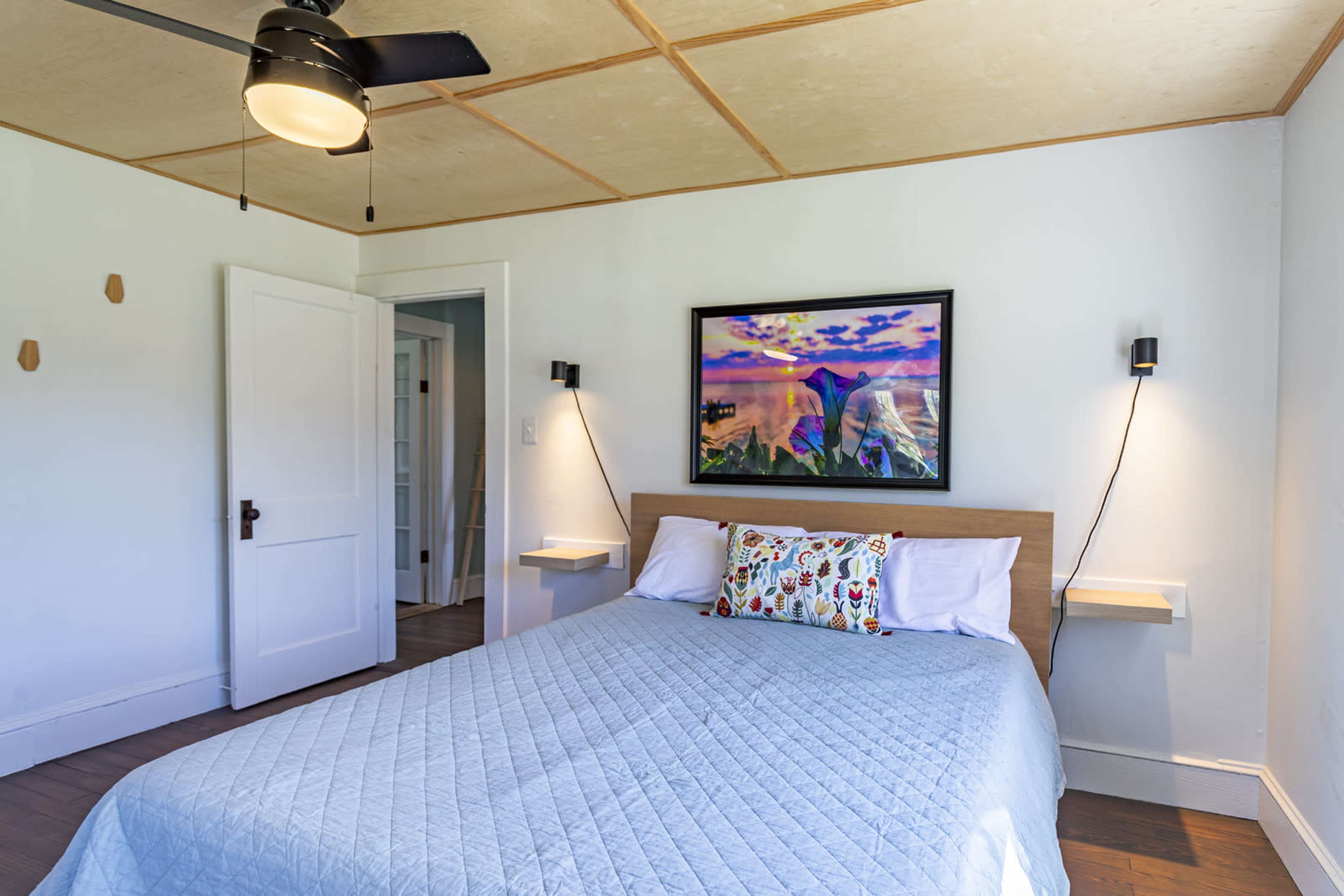 A well-lit bedroom features a bed with a patterned pillow, a framed landscape painting on the wall, and simple wall sconces on either side.