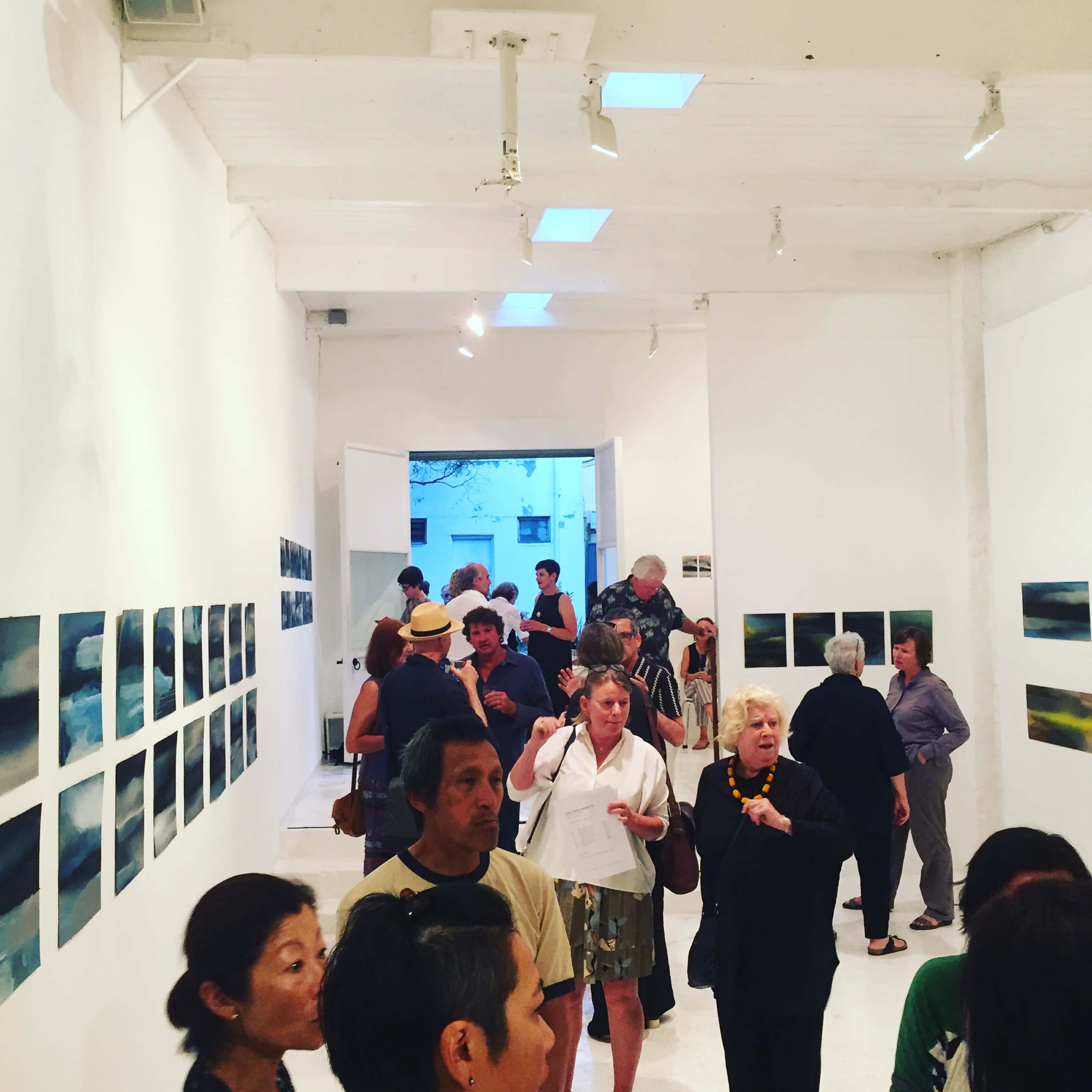 The image shows a crowded art gallery featuring people engaged in conversation while examining a series of paintings displayed on the white walls.