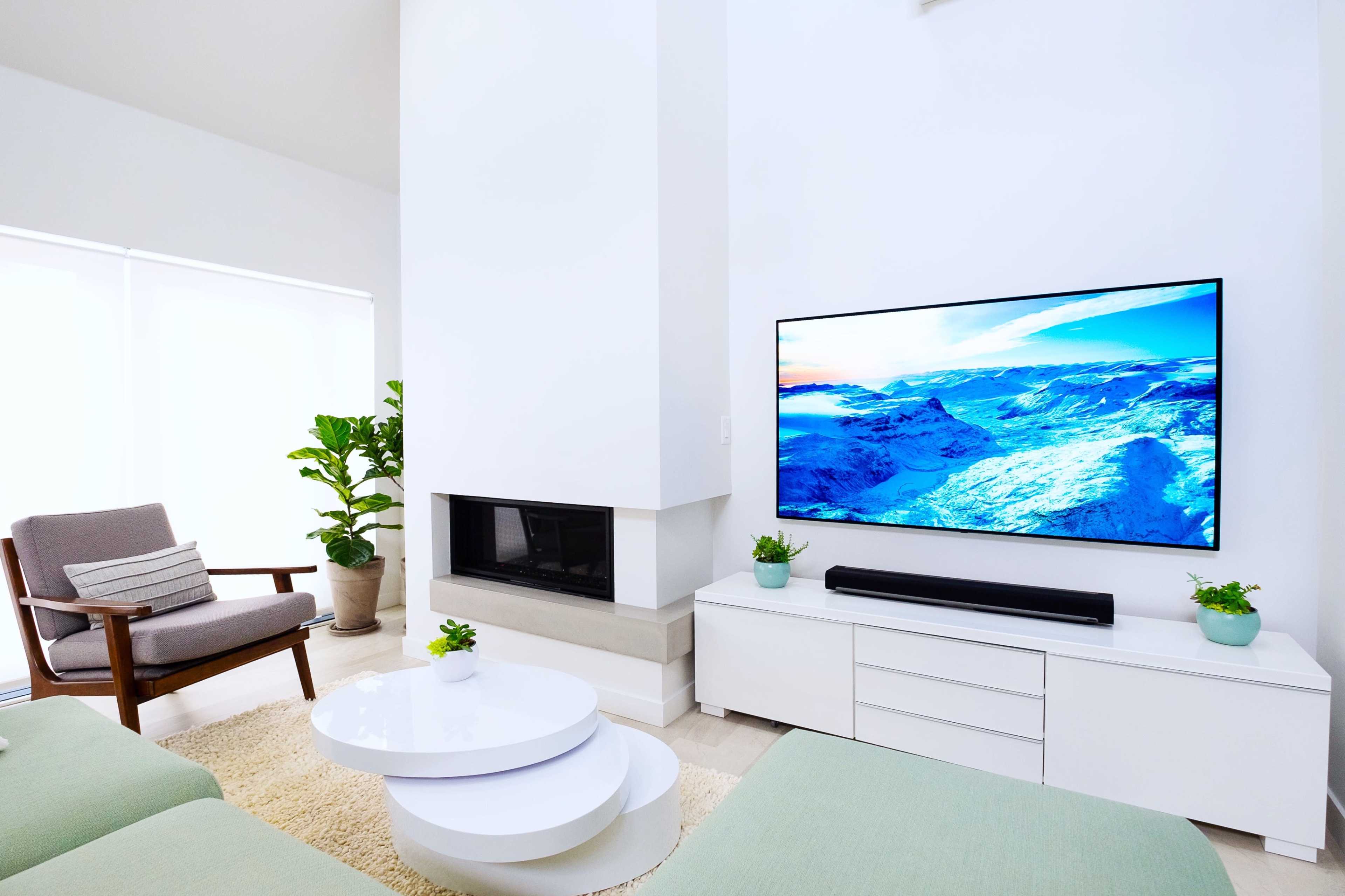 A modern living room with a large flat-screen TV mounted on a white wall, a white media console, a circular coffee table, and a comfortable armchair beside a potted plant.