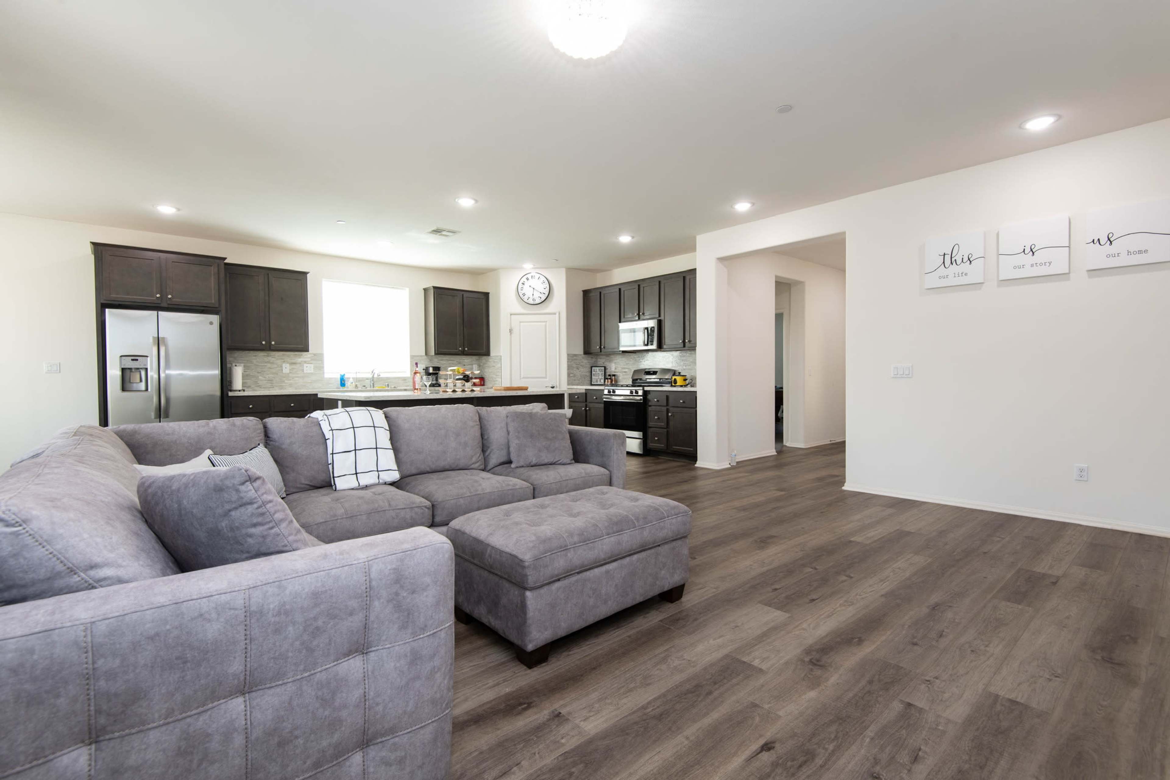 A modern living room features a gray sectional sofa, hardwood floors, and an open kitchen with dark cabinets and stainless steel appliances.