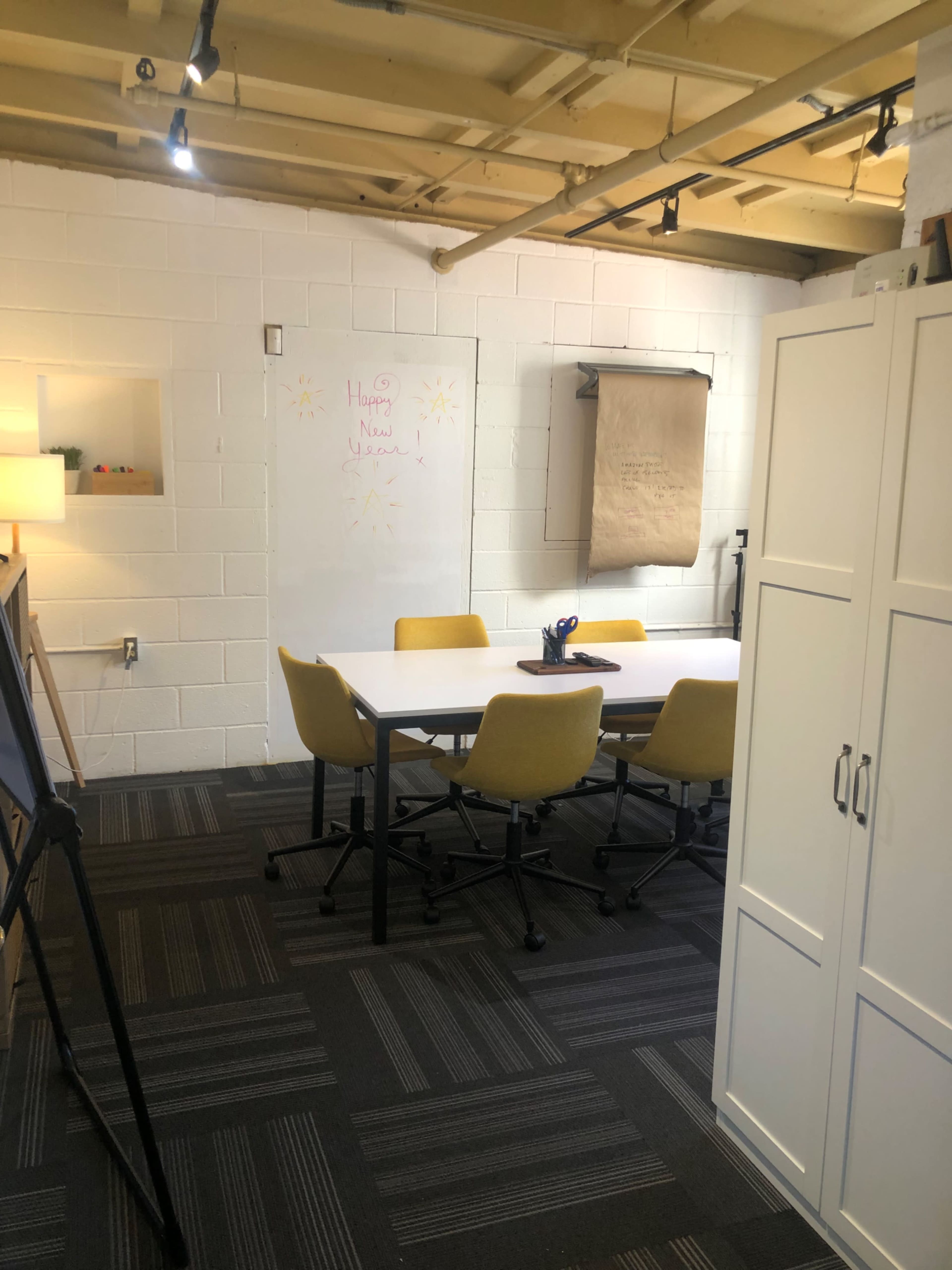 A small conference room with a whiteboard, a large table surrounded by yellow chairs, and a storage cabinet against the wall.