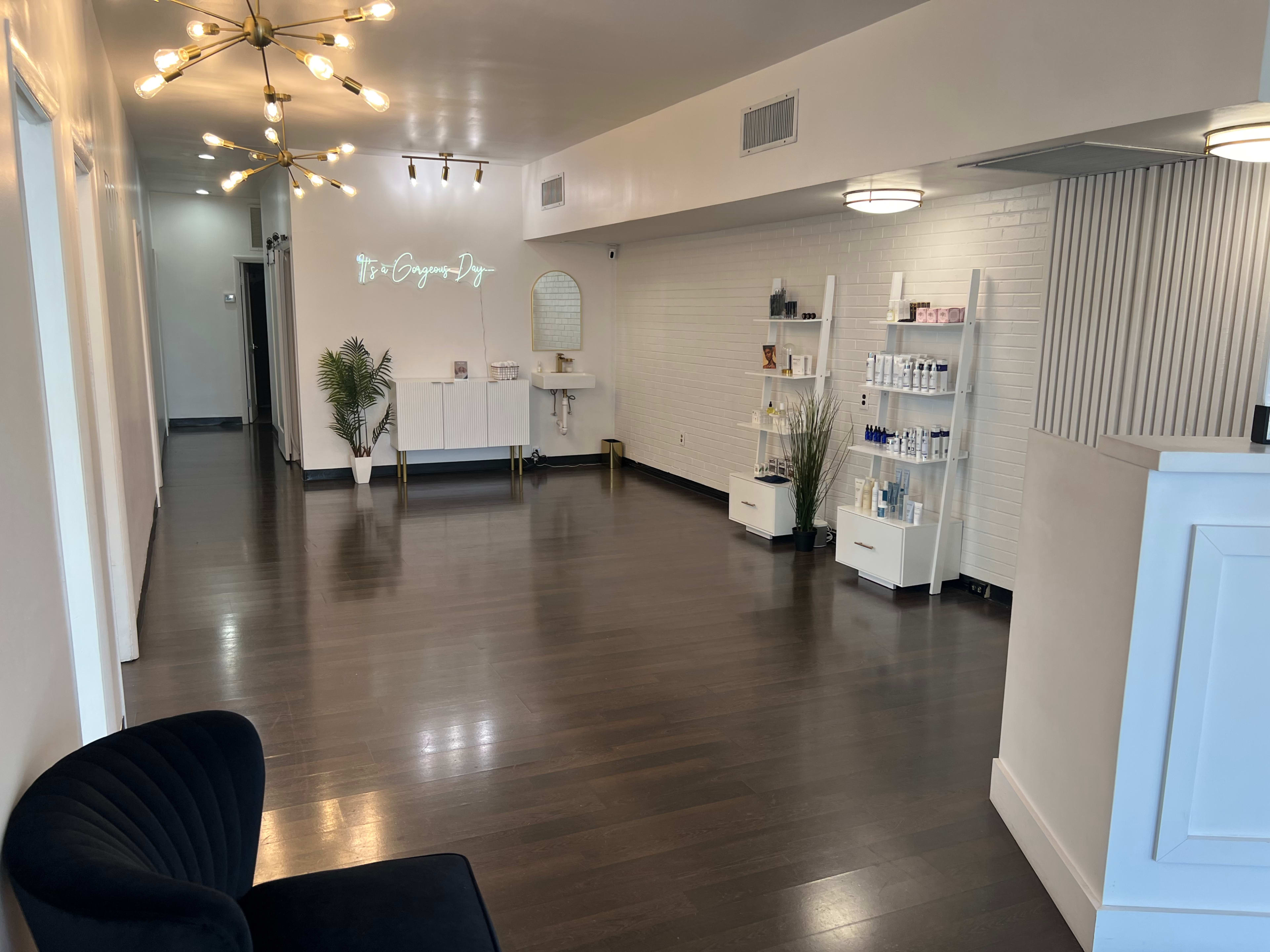 A modern, spacious reception area of a beauty or wellness clinic, featuring a central lighting fixture, white walls, and shelves displaying various products.