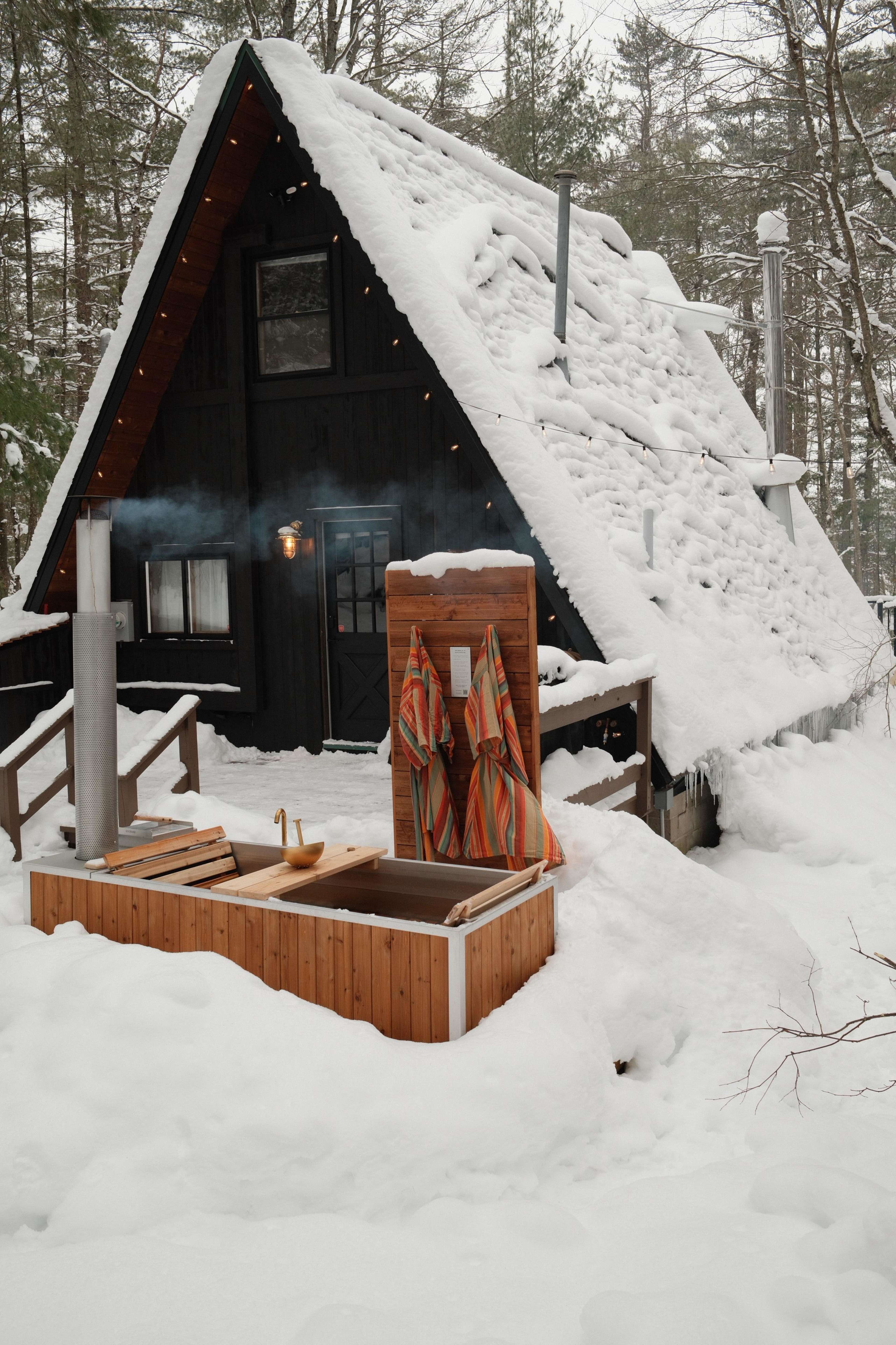 A black A-frame cabin with a snowy roof is situated in a forested area, featuring a wooden hot tub and two colorful towels hanging nearby.