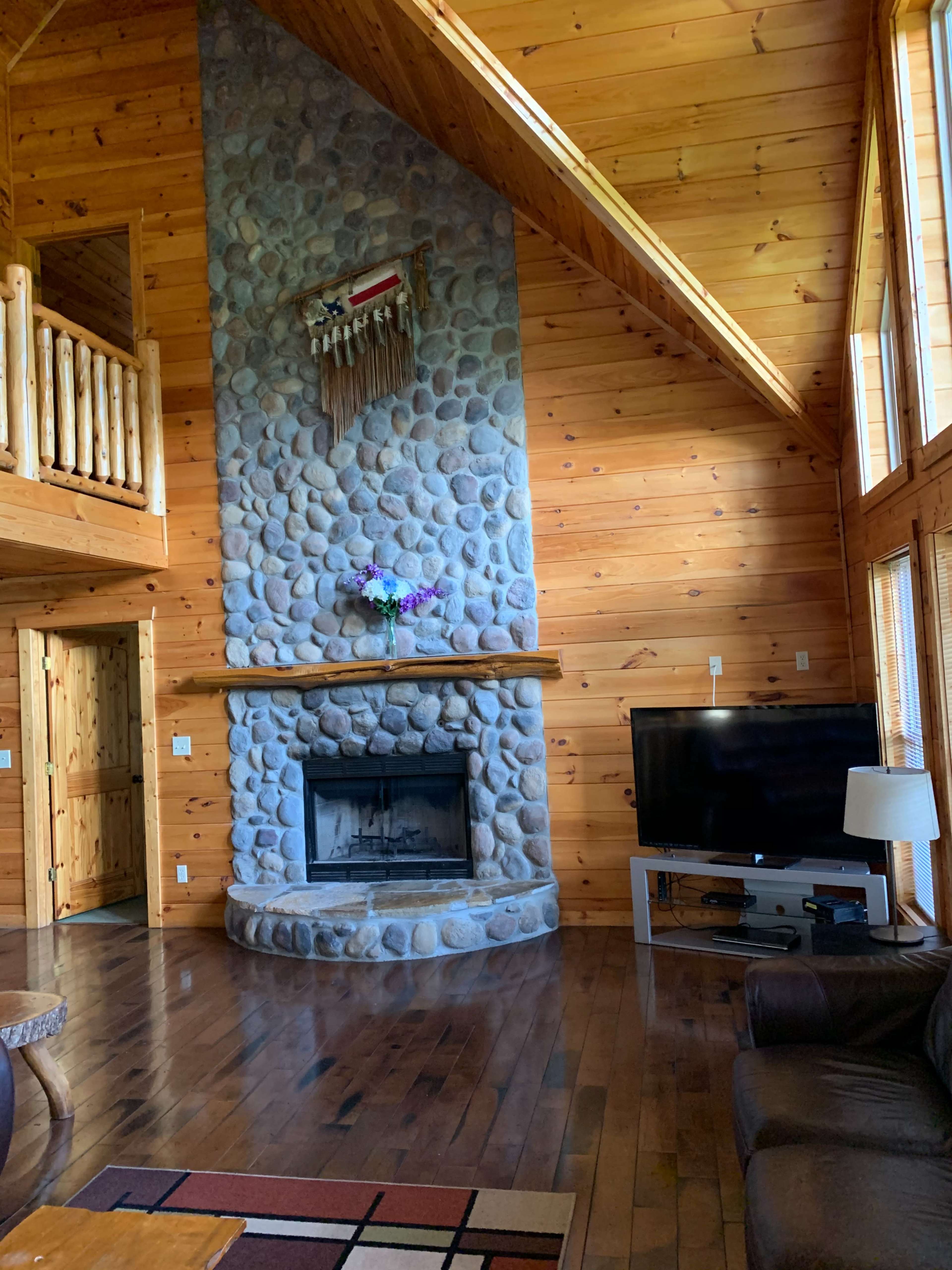 A spacious wooden living room with a stone fireplace at its center, large windows, and a television on a white console.