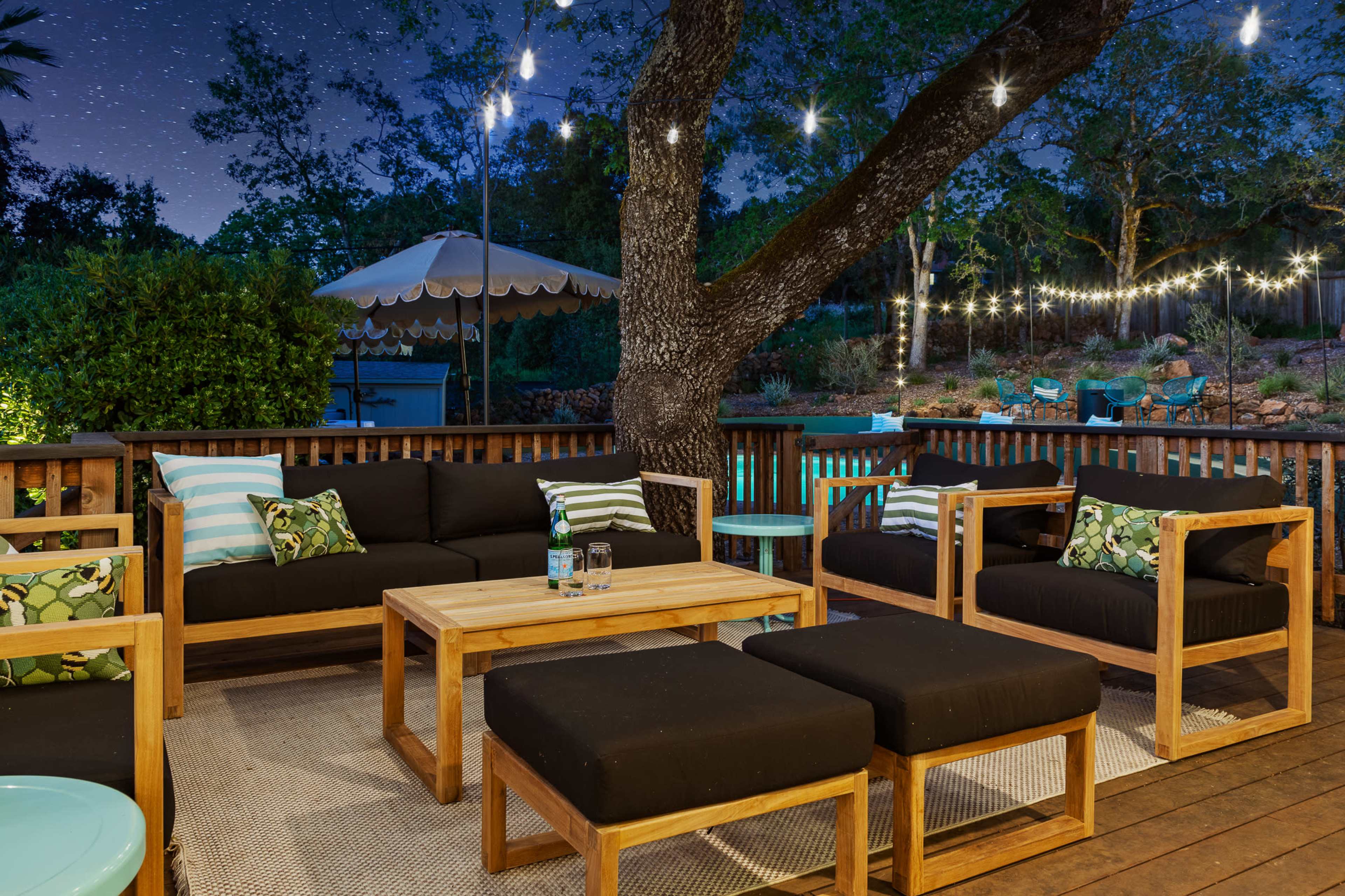 A wooden patio area features dark seating, a central table, and string lights illuminating a nearby tree and swimming pool in the background.