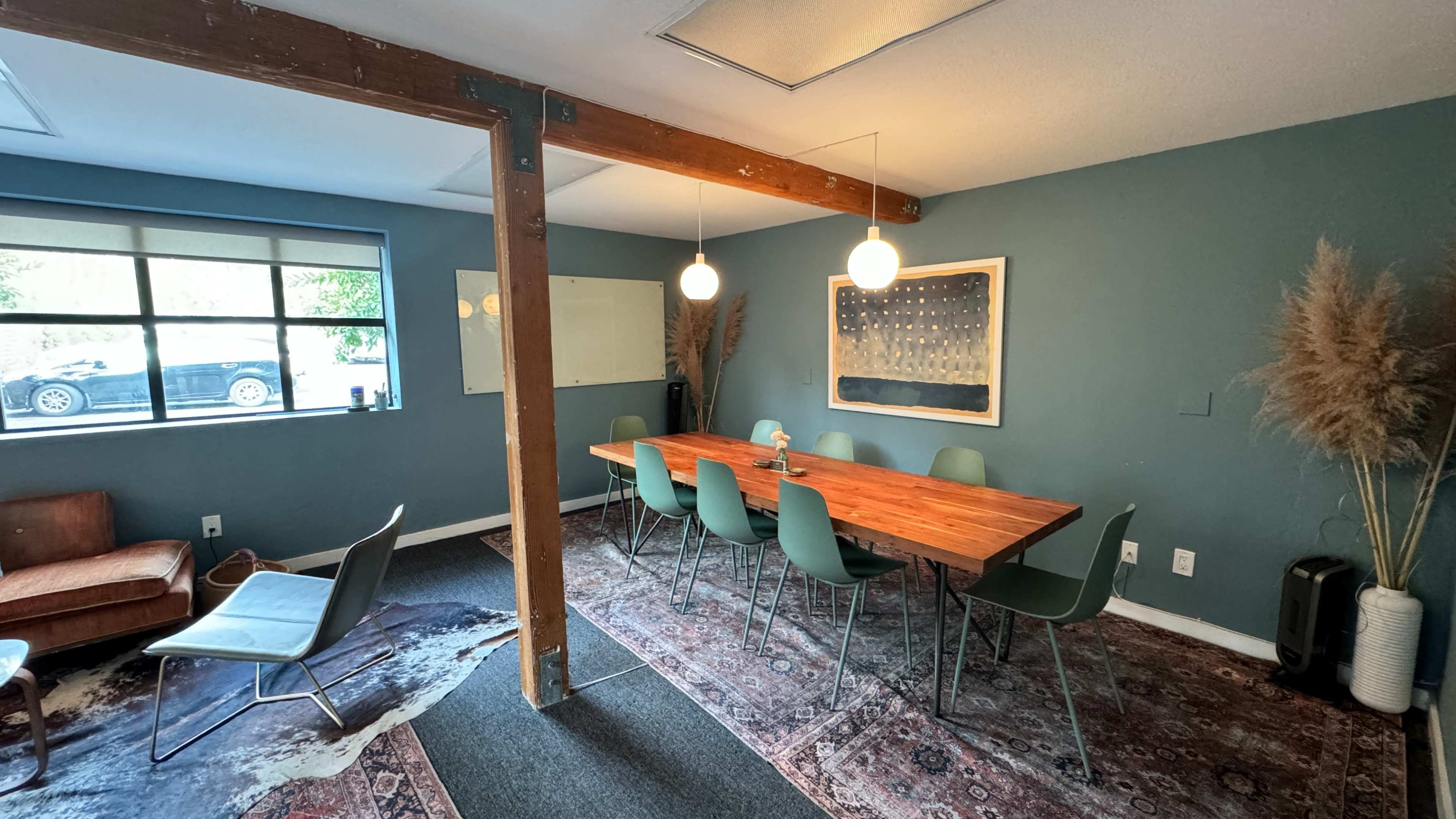 A conference room features a long wooden table surrounded by green chairs, with a large wall art piece, a whiteboard, and a window showing a parked car outside.
