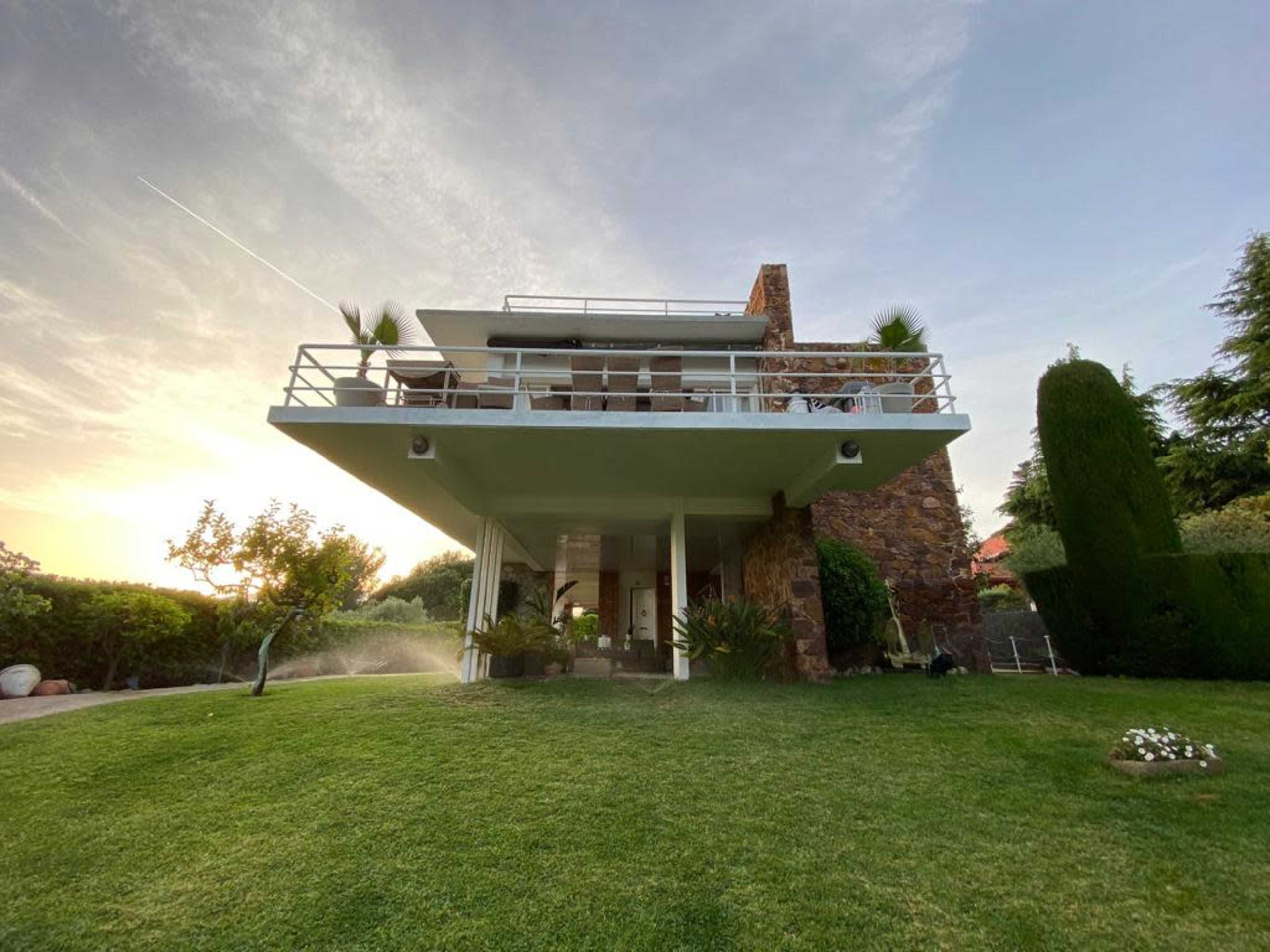 A modern two-story house with a flat roof and a stone wall, surrounded by greenery and stacked balconies.