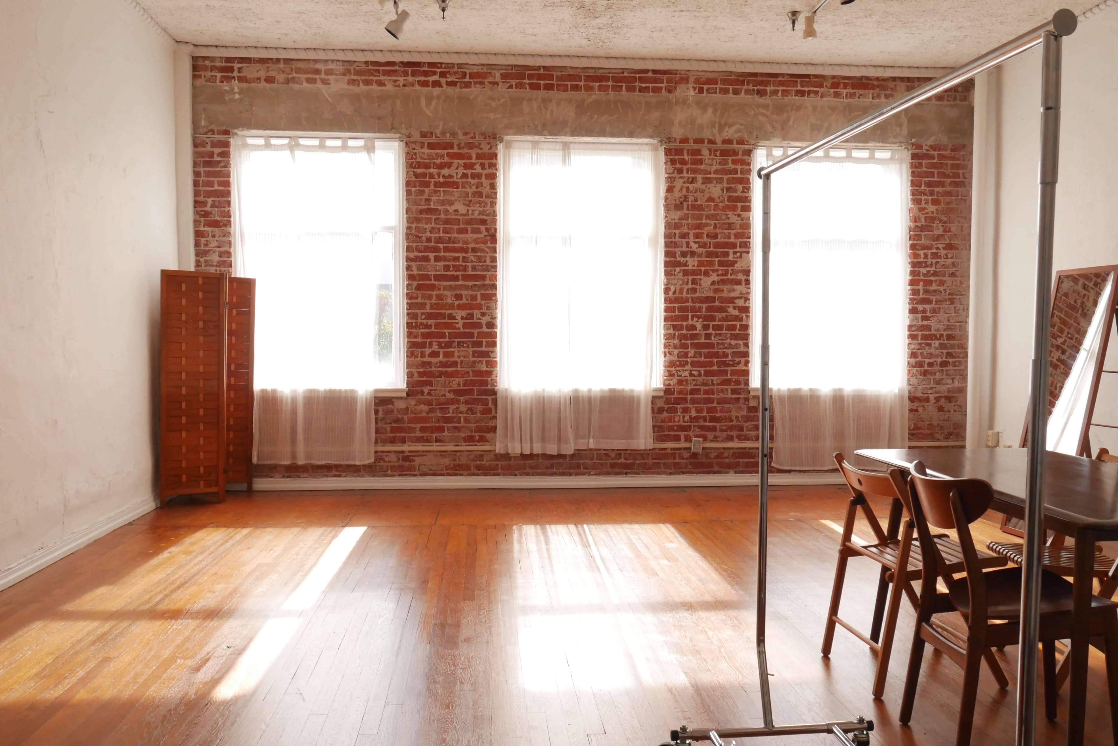 A spacious room with exposed brick walls, large windows allowing sunlight to enter, and a set of wooden chairs beside a clothing rack.