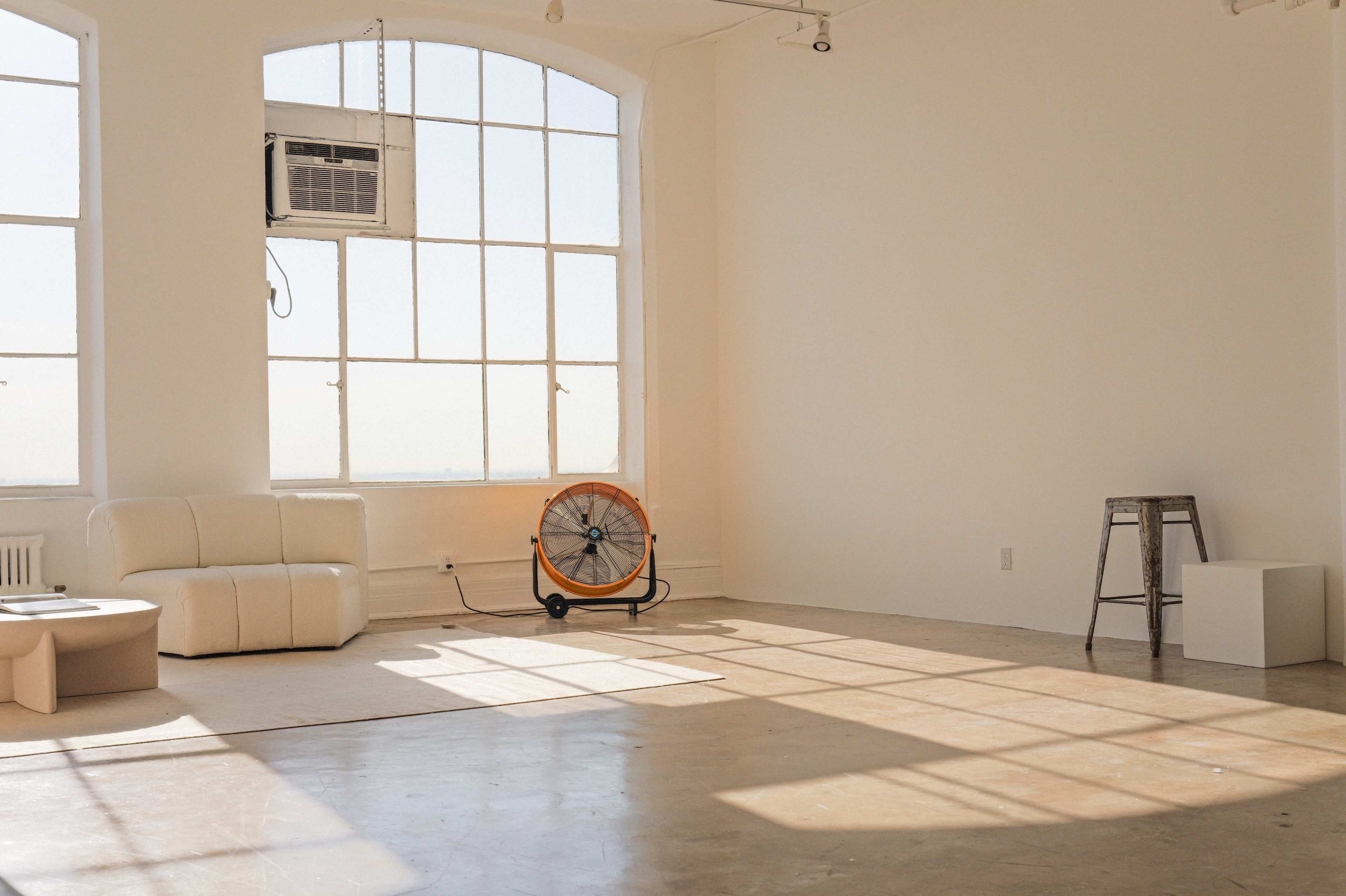The image shows a bright, empty room with large windows, a white couch, an industrial fan, a folding stool, and a small cube-shaped table.