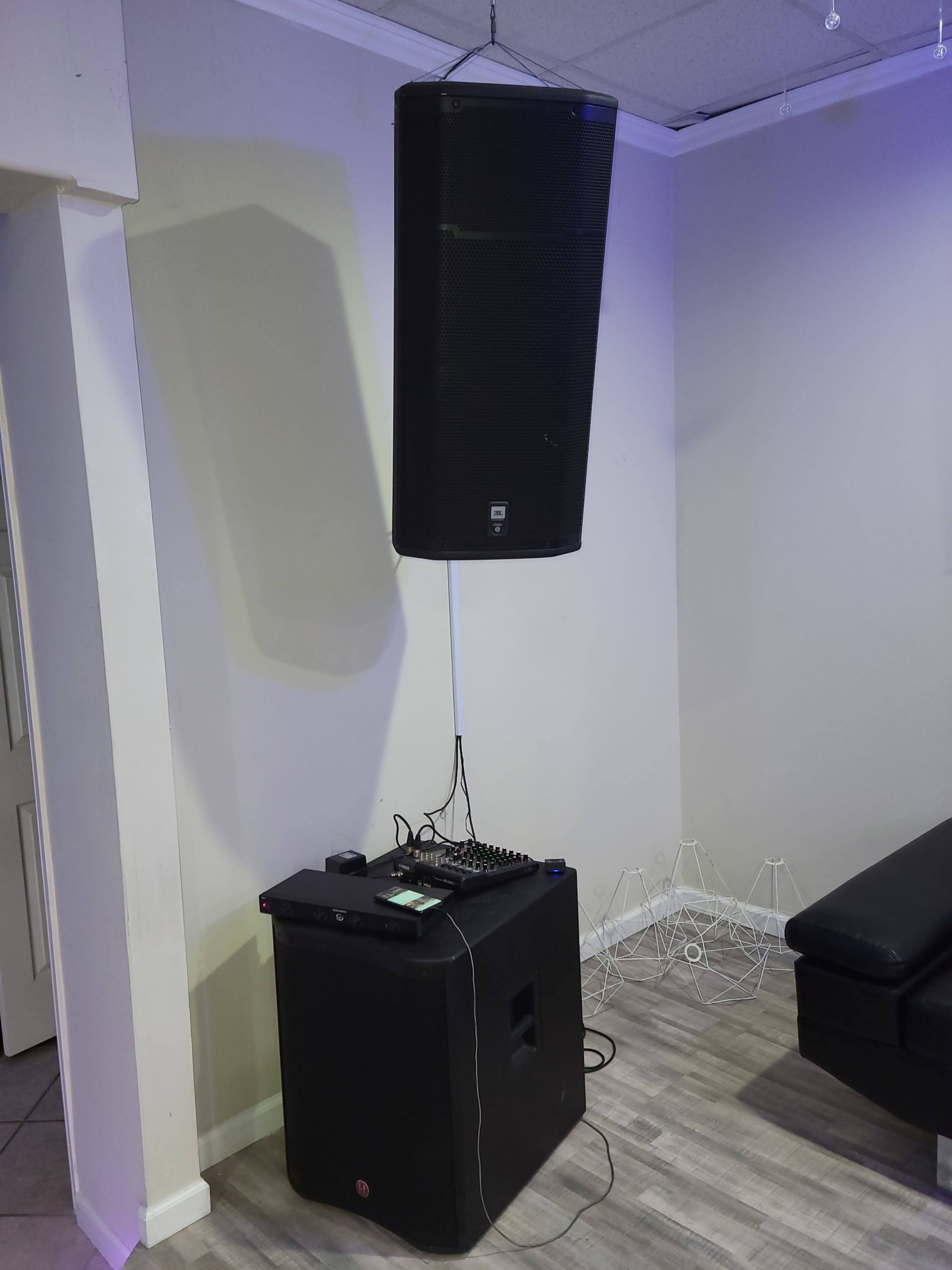 A large speaker is suspended from the ceiling above a smaller speaker and a DJ controller on a table in a room with light-colored walls and flooring.