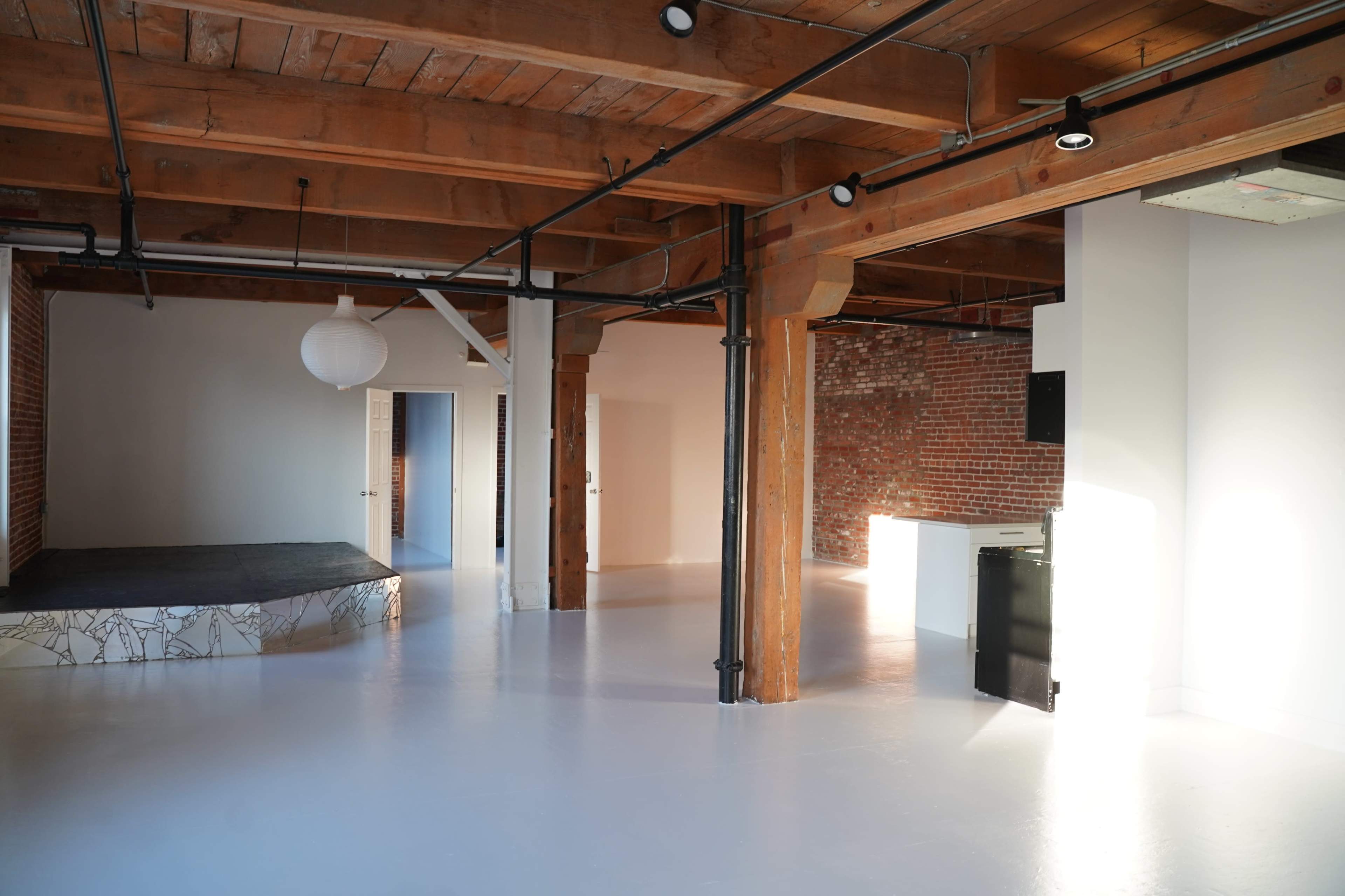 The image shows a spacious, empty room with wooden beams and columns, featuring a mix of brick and smooth white walls, and an area with a black platform.
