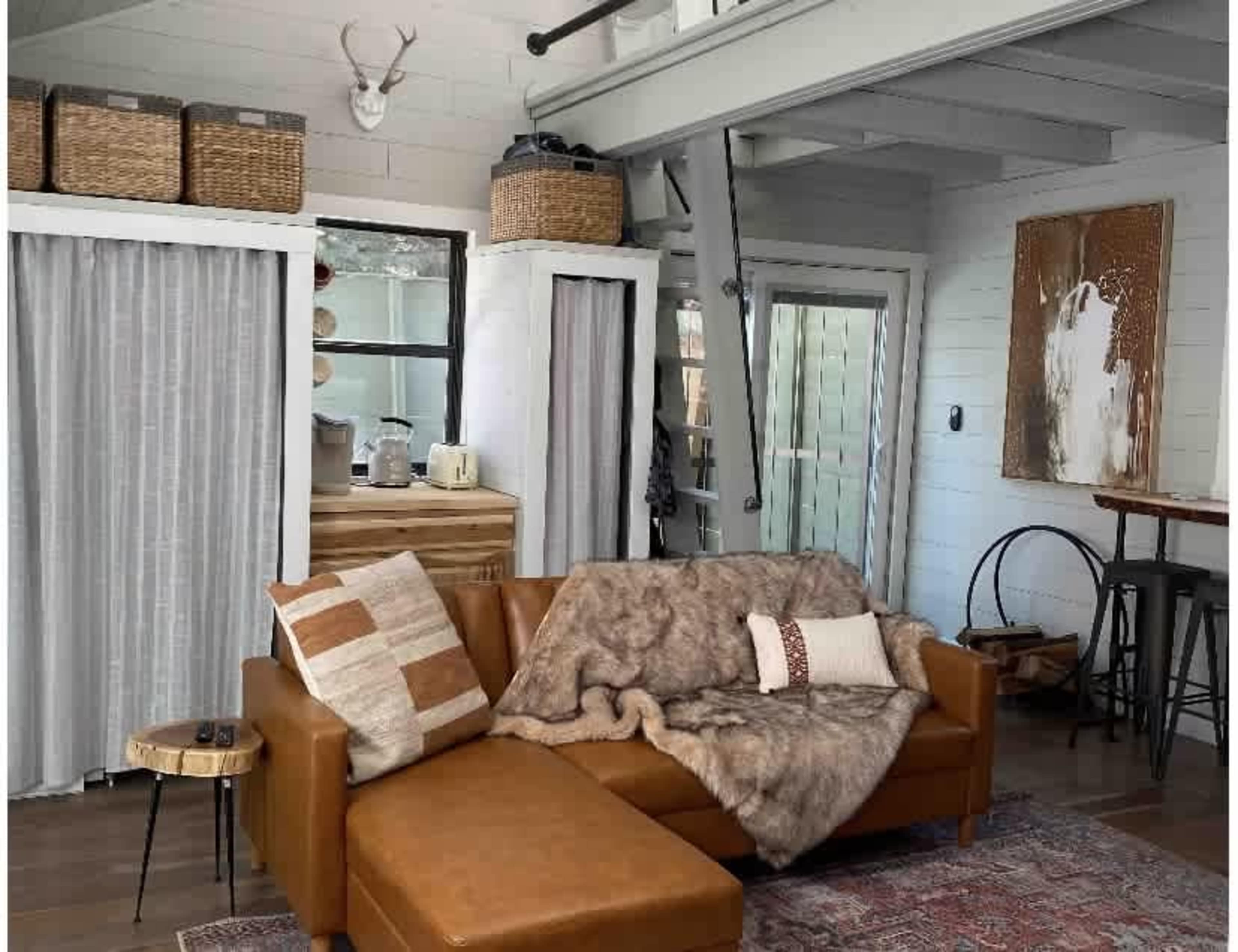 A cozy living room with a brown leather sectional couch, a fur throw blanket, a small round side table, and a rustic wooden cabinet against white-paneled walls.