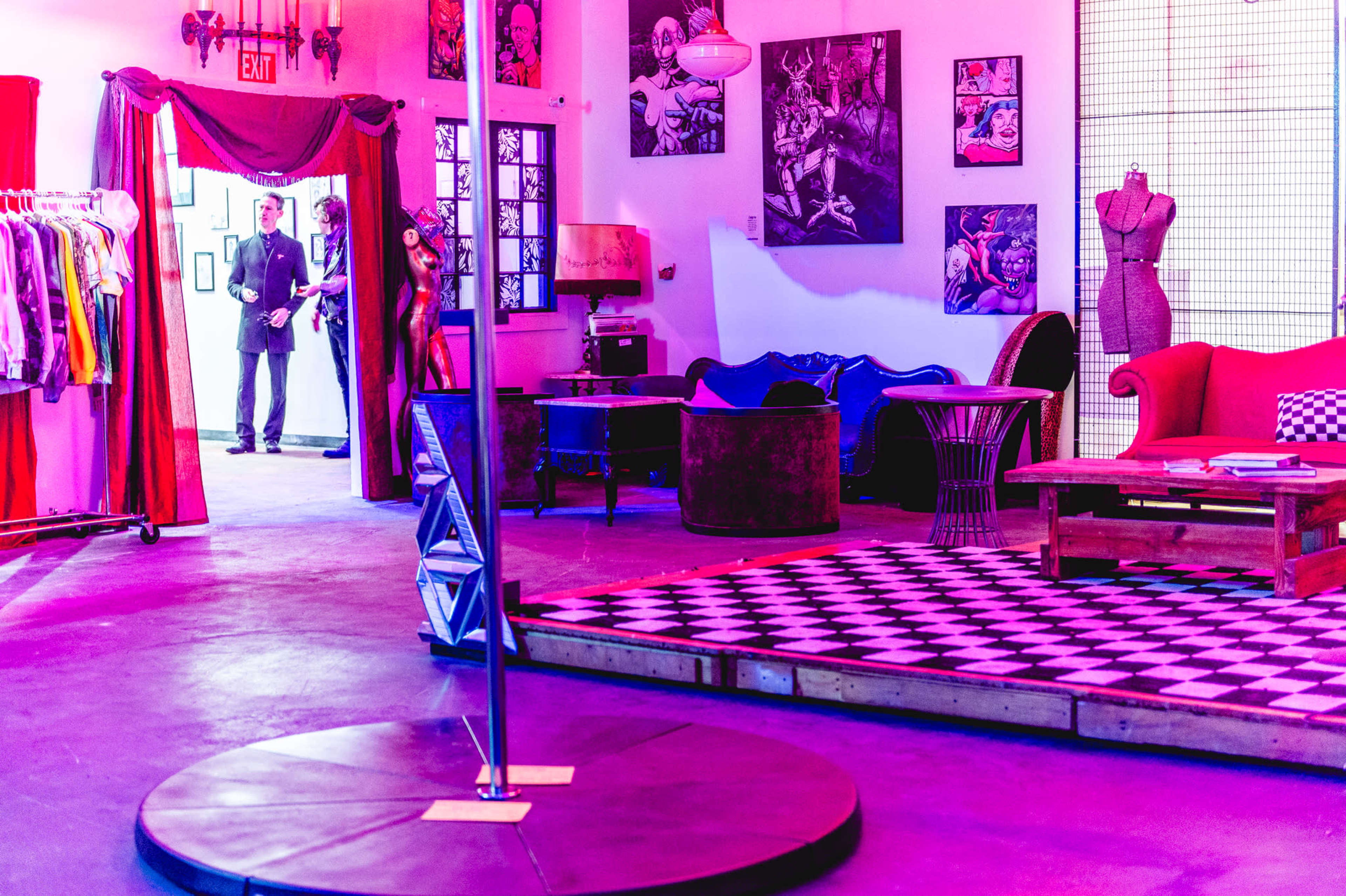 A vibrant room featuring a pole in the center, surrounding seating areas, clothing racks, and artistic wall decor.