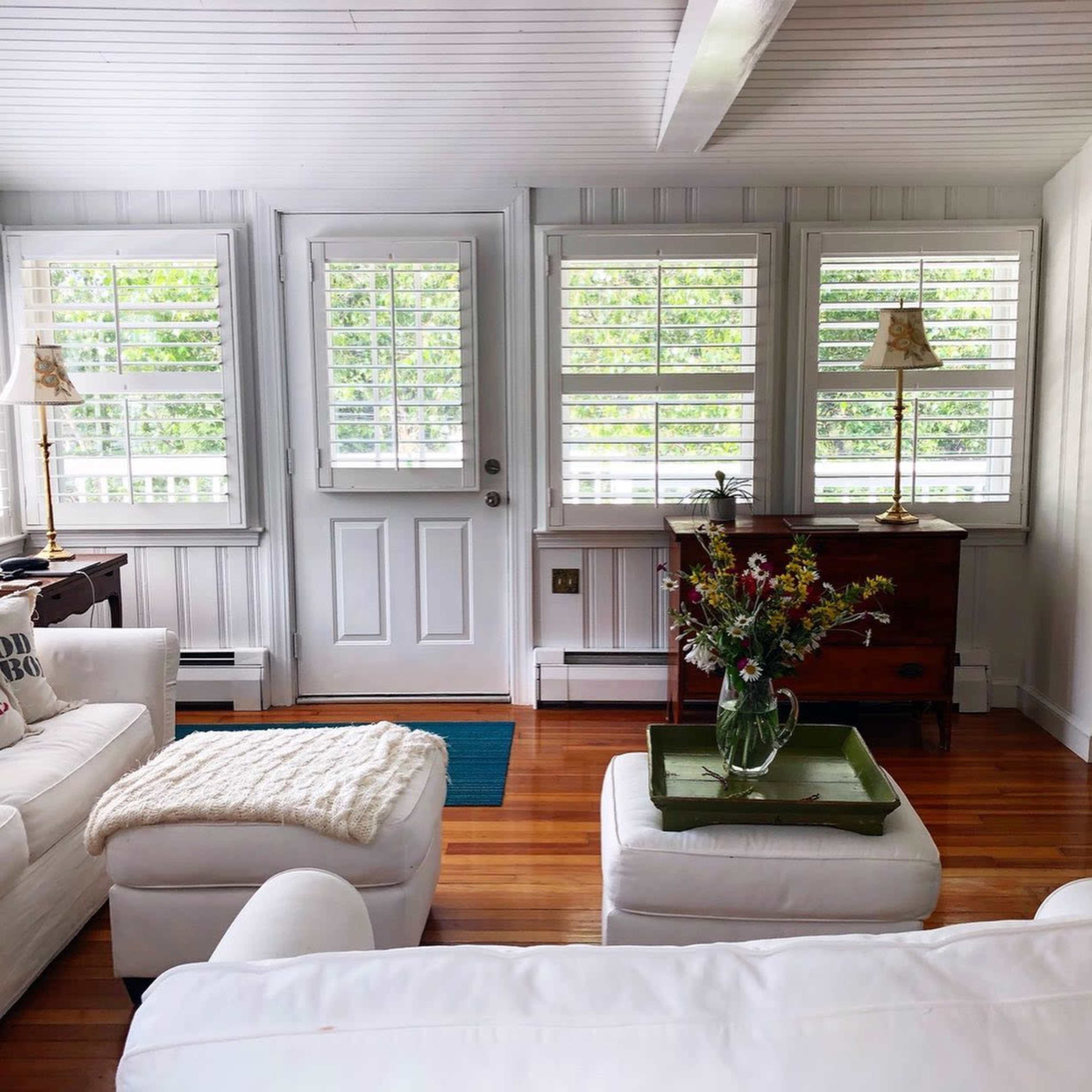 The room features white furniture, large windows with plantation shutters, and a wooden door leading outside, complemented by a wooden table and a vase of flowers on a green tray.
