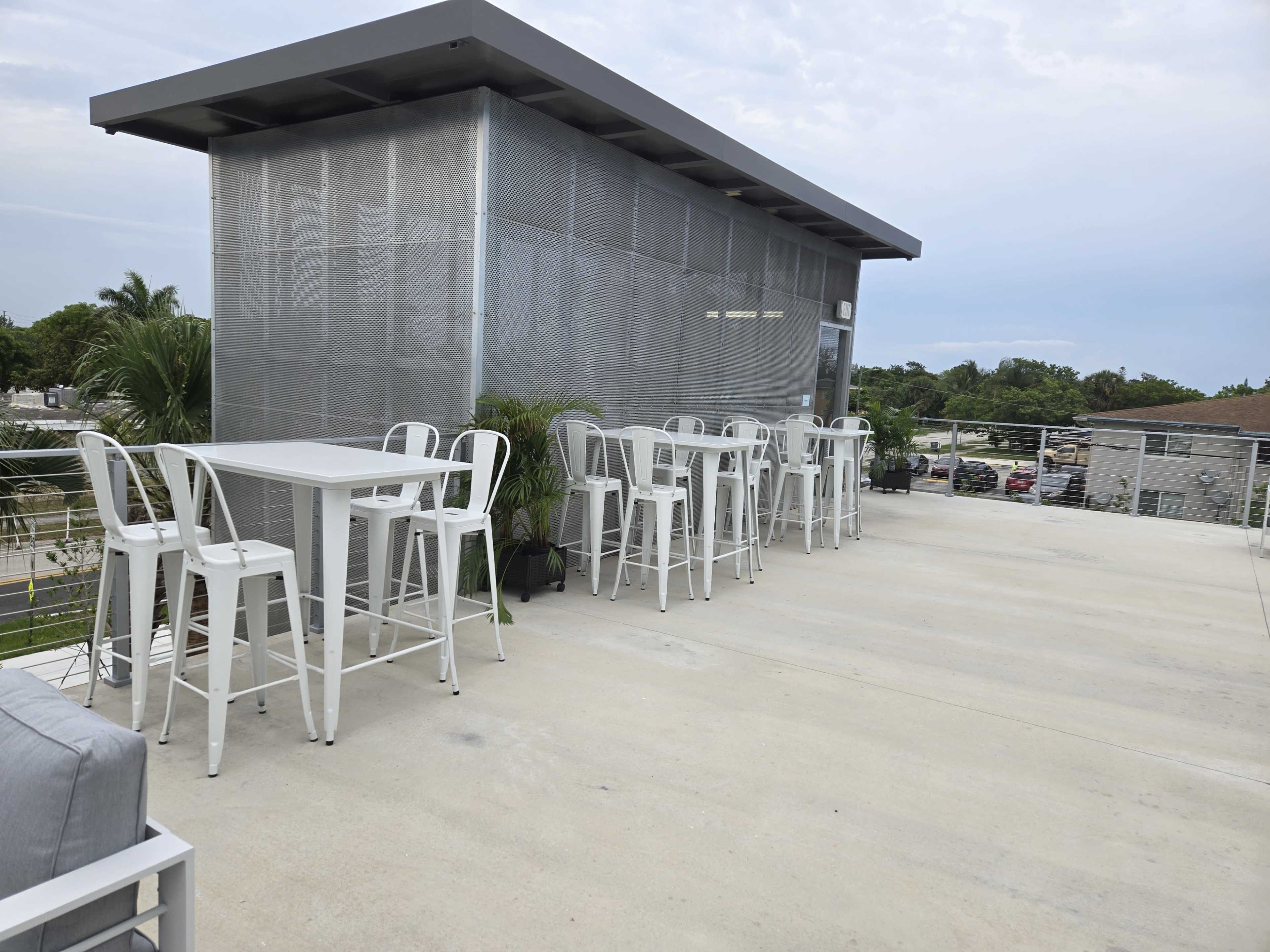A rooftop patio features a series of white metal tables and stools next to a modern structure surrounded by palm trees.