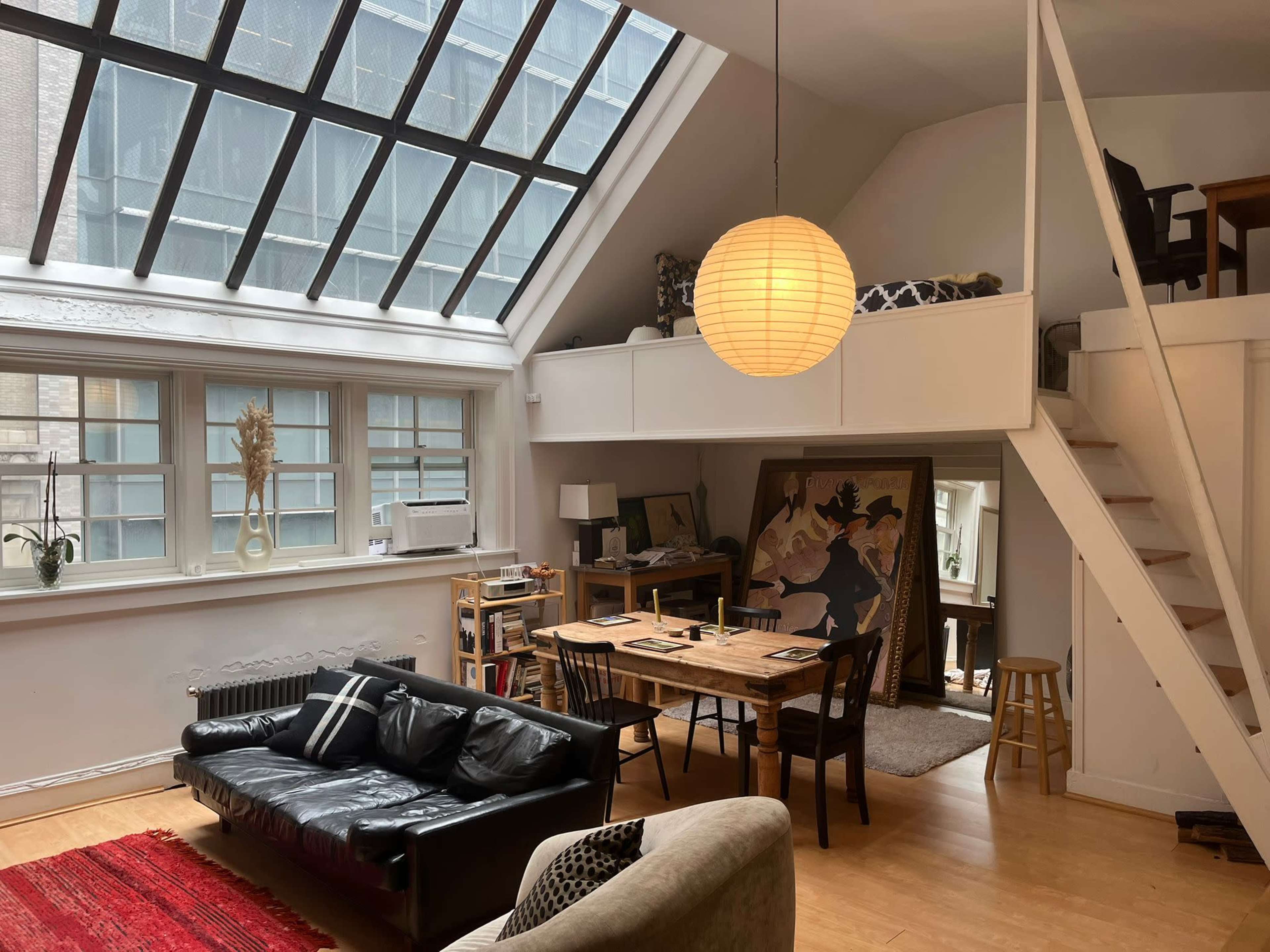 The image shows a modern loft-style living space with a high ceiling, large windows, a black leather sofa, a dining table, and a staircase leading to a loft area.