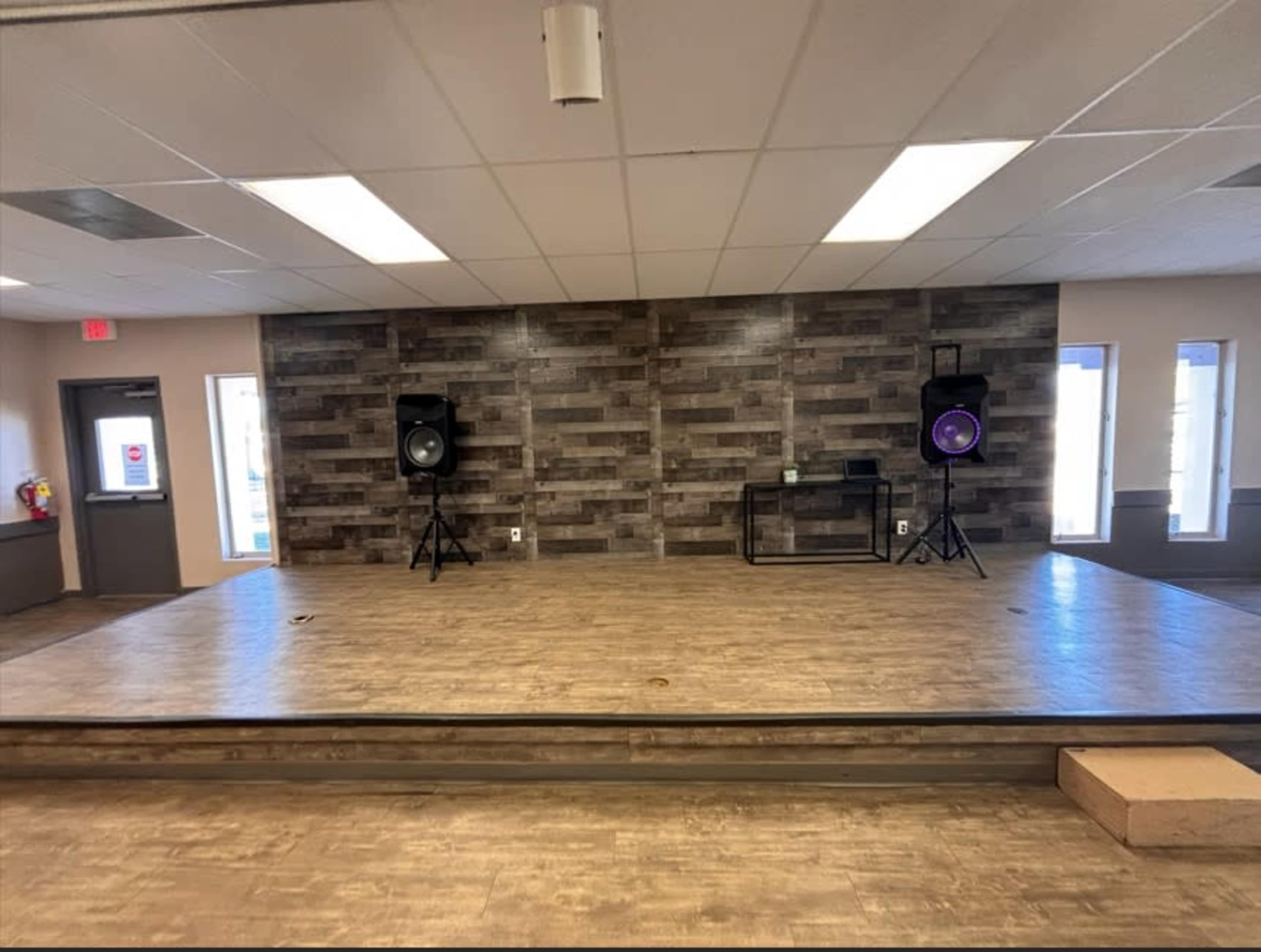 The image shows a stage area with wooden flooring, flanked by two speakers on stands, and a textured wall in the background.