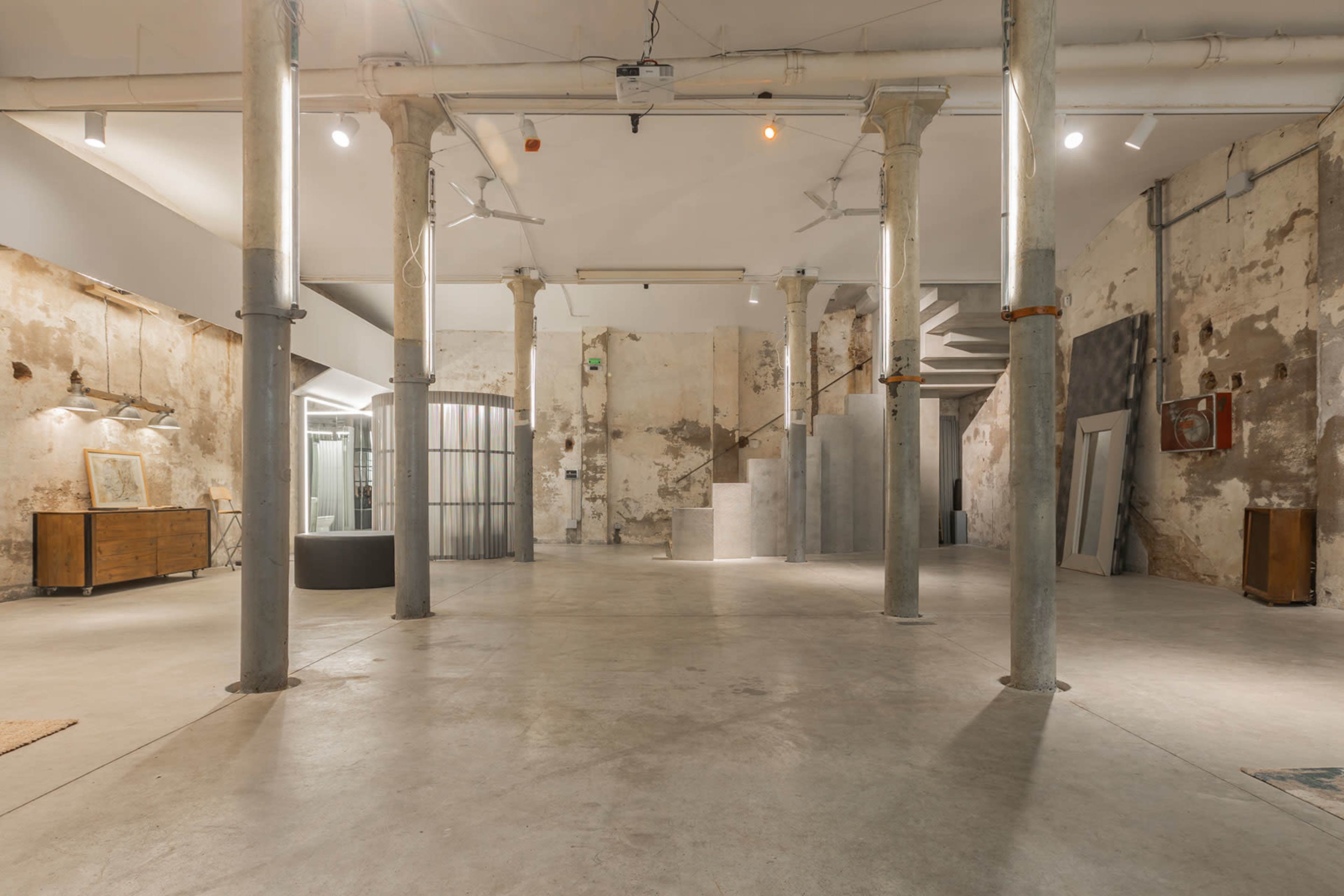 The image shows an industrial-style interior space with exposed concrete walls, tall pillars, and scattered furniture in a well-lit area.