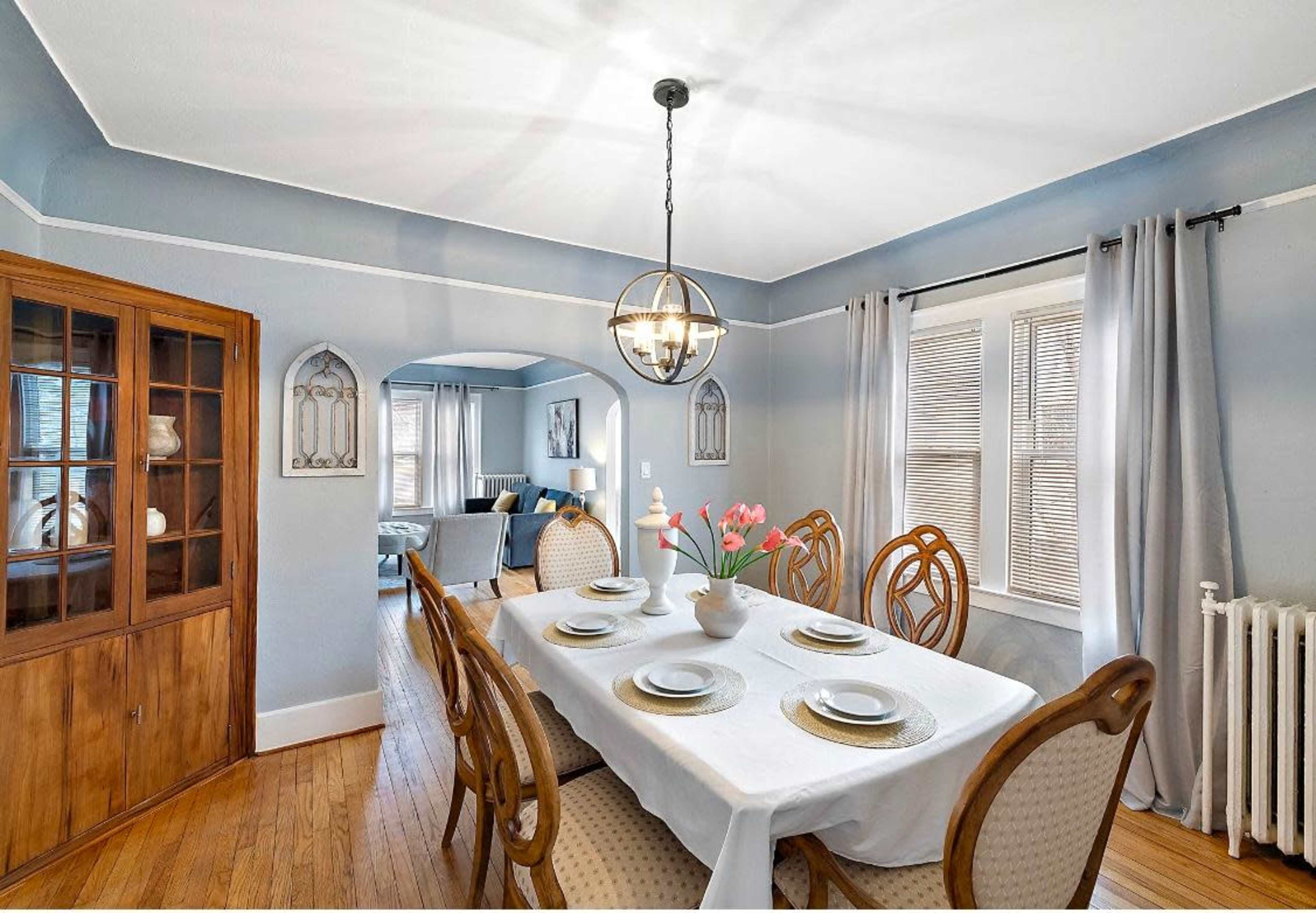 A dining room features a large table set for six, with a vase of flowers at the center, surrounded by light blue walls and wooden accents.