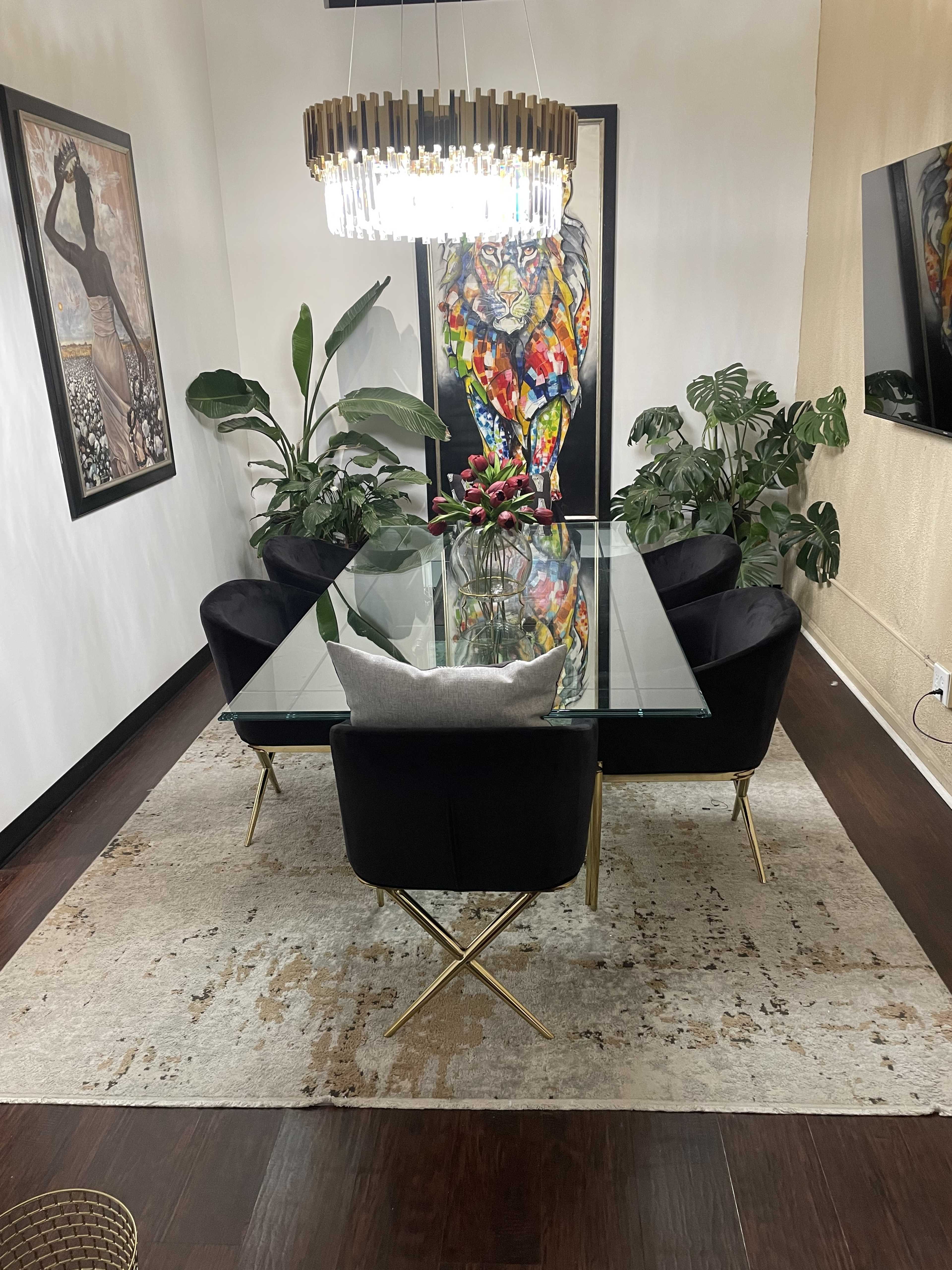 A modern dining room with a glass table surrounded by black velvet chairs, a large colorful lion painting on the wall, and indoor plants on either side.