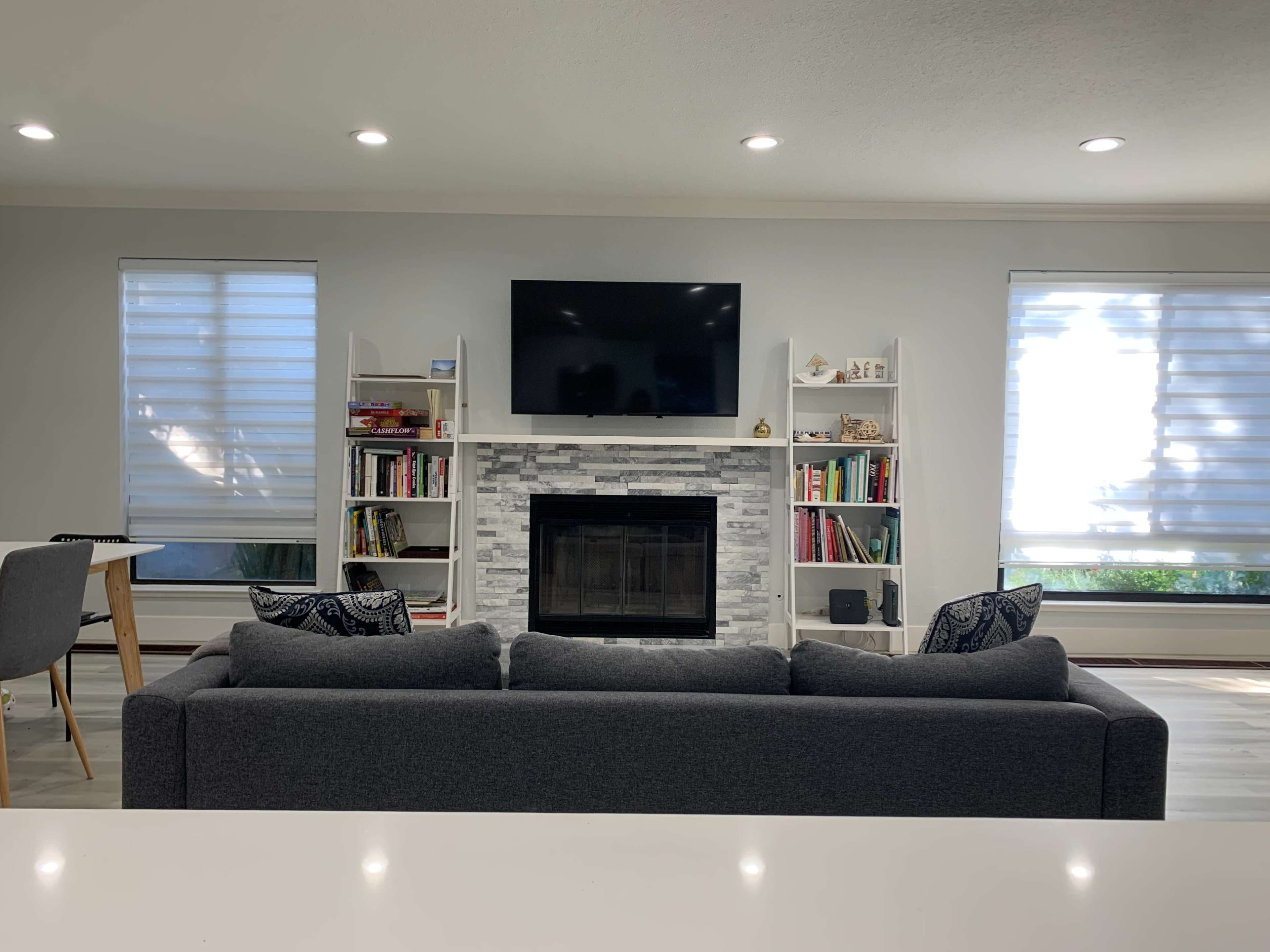 A modern living room with a gray couch facing a stone fireplace and a wall-mounted television, surrounded by bookshelves and large windows with shades.