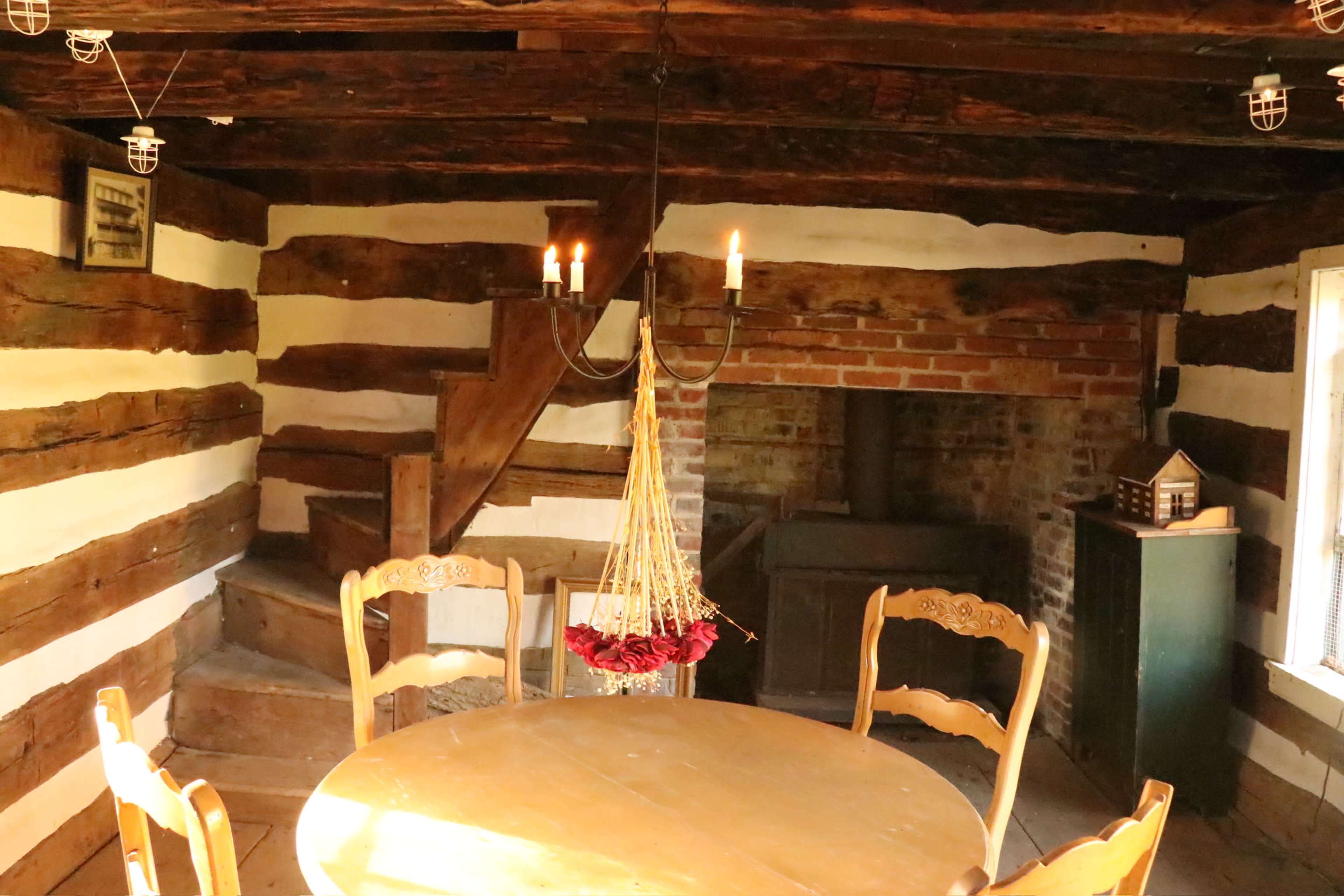 The image shows a rustic interior of a cabin with wooden beams, a round wooden table, chairs, a chandelier, and a staircase leading to an upper level.