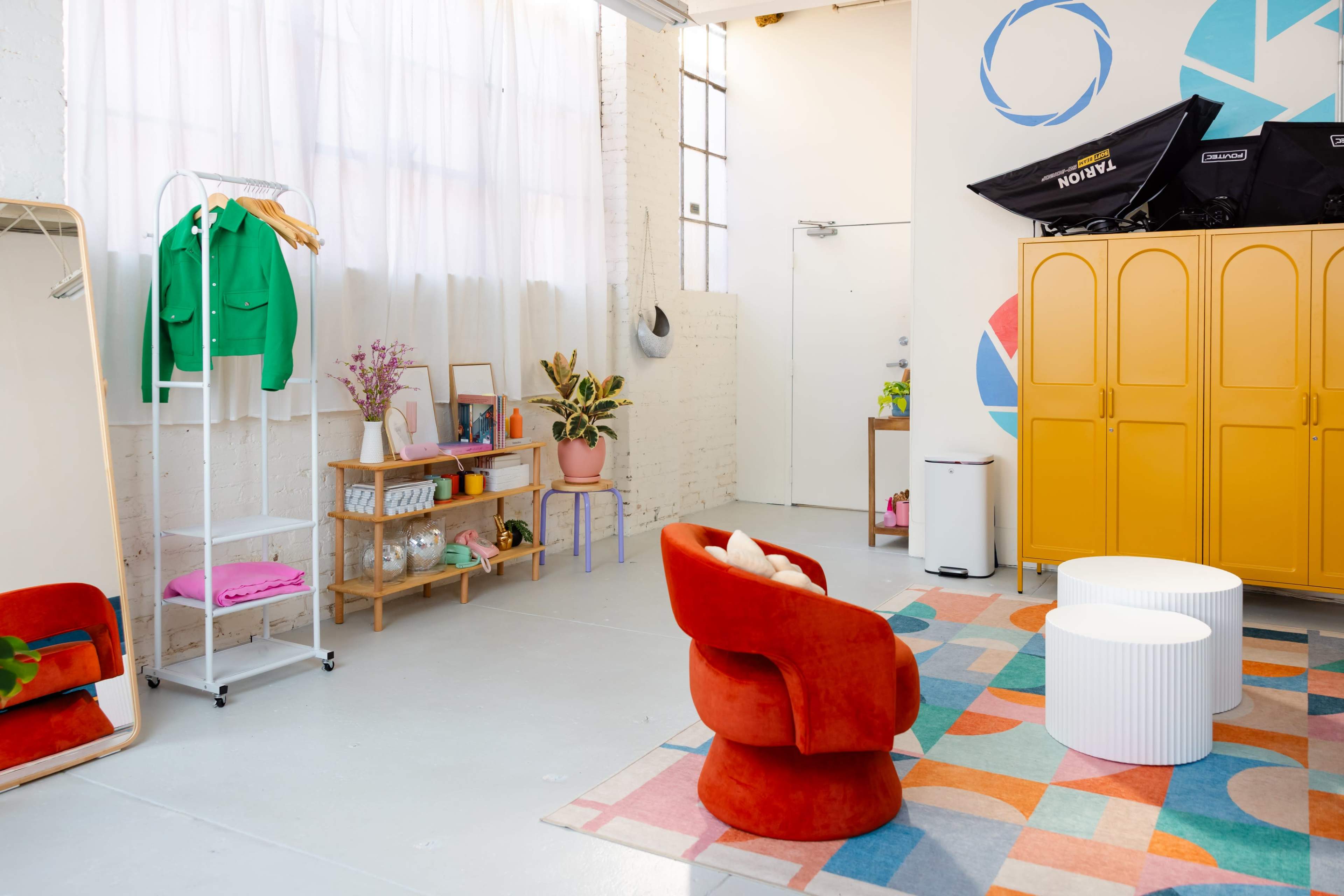 The image shows a brightly lit room with a large mirror, a yellow cabinet, colorful furniture, a rug with geometric patterns, and decorative plants.