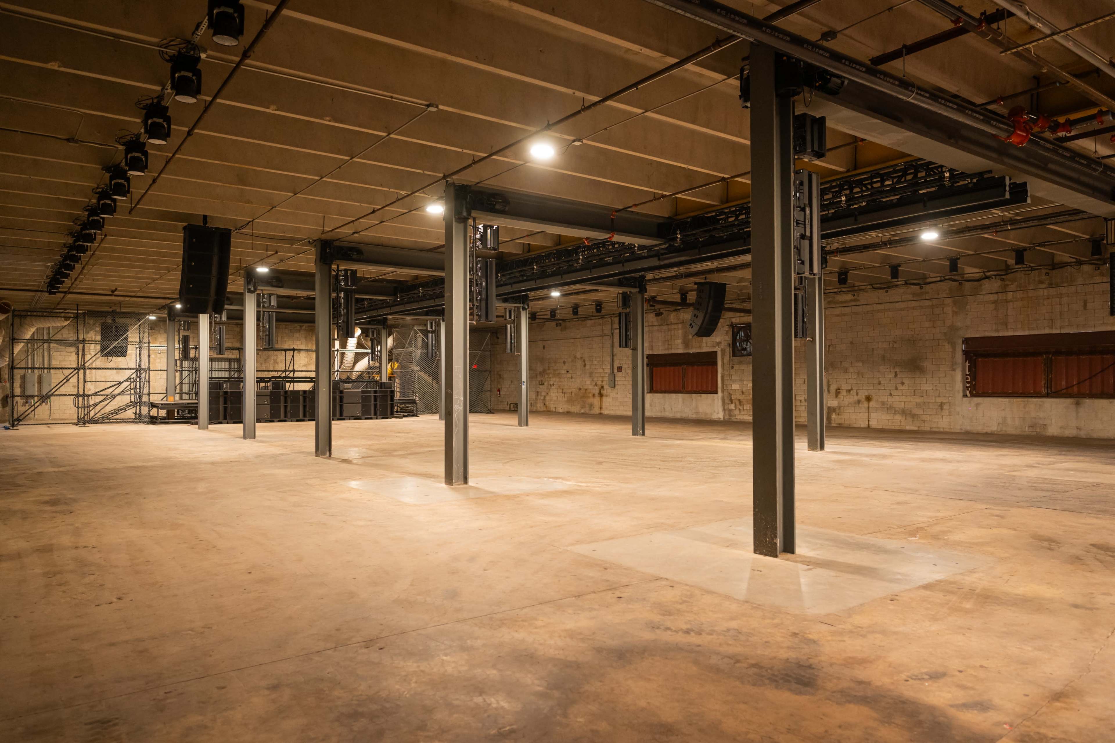 The image shows an empty industrial space with concrete floors, metal support beams, and overhead lighting, designed for events or performances.