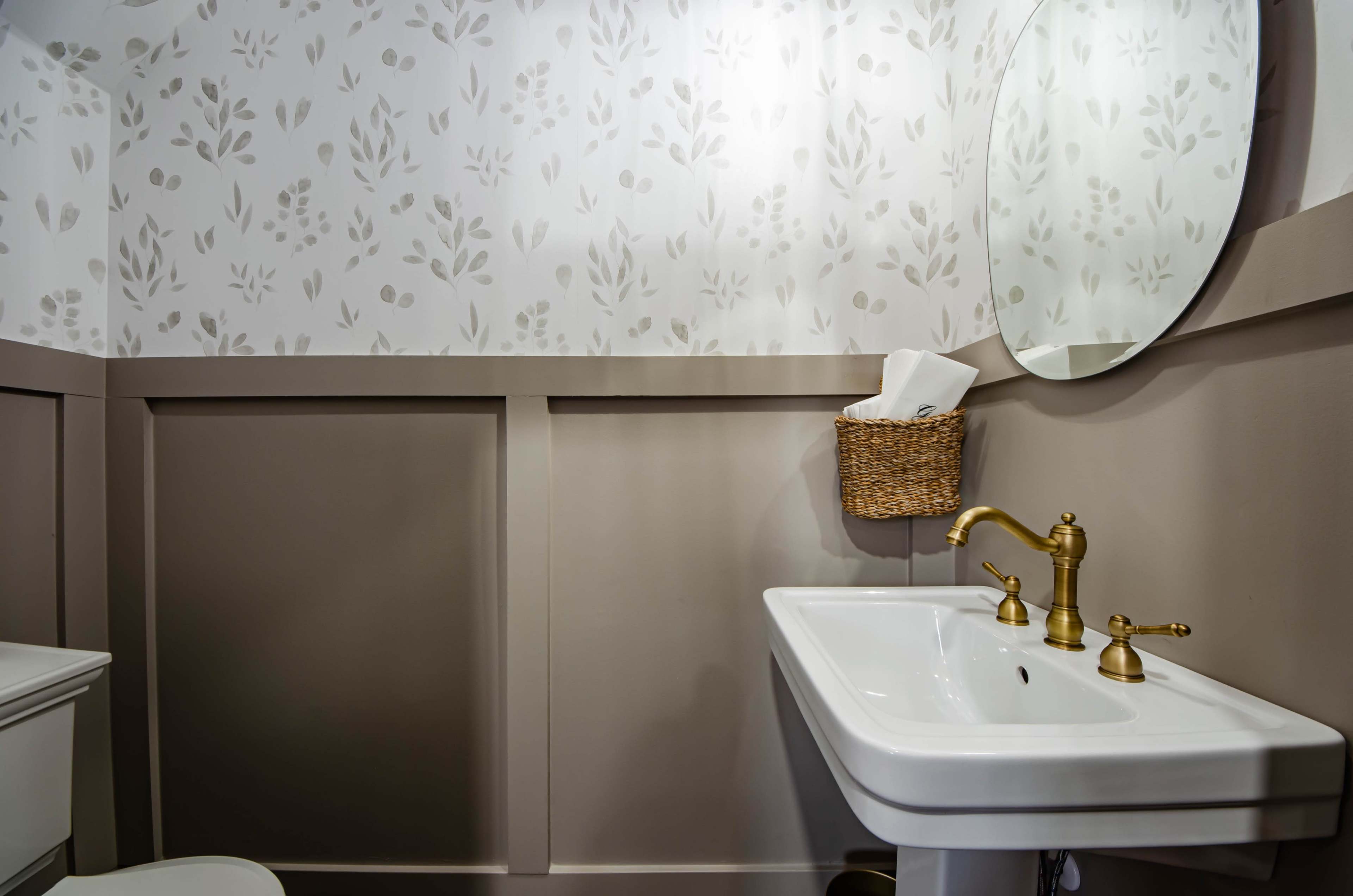 A bathroom with a white sink, gold faucet fixtures, a round mirror, and floral wallpaper above a beige wainscoting.
