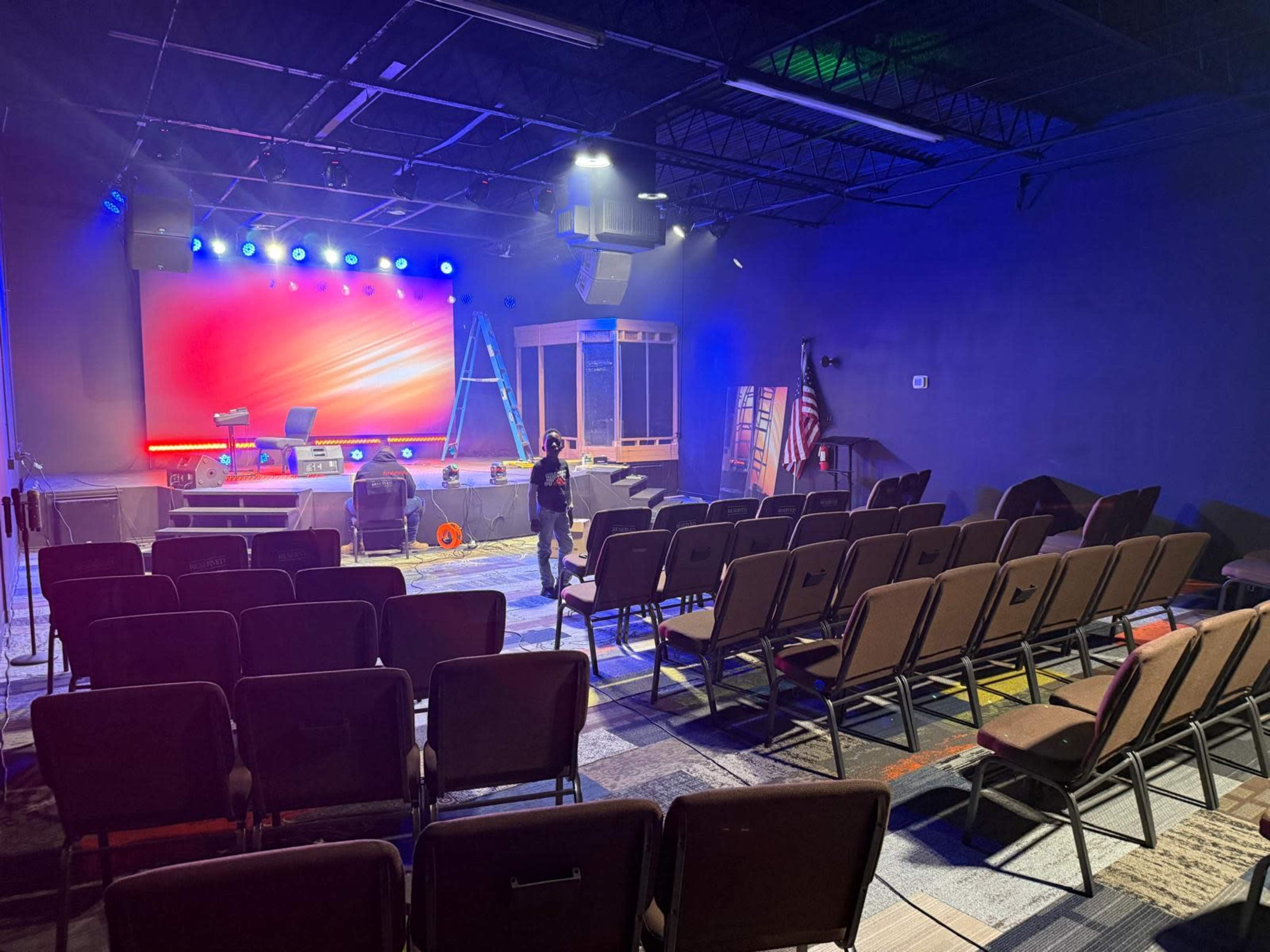 A spacious auditorium features rows of chairs facing a brightly lit stage with a large backdrop and a ladder nearby.