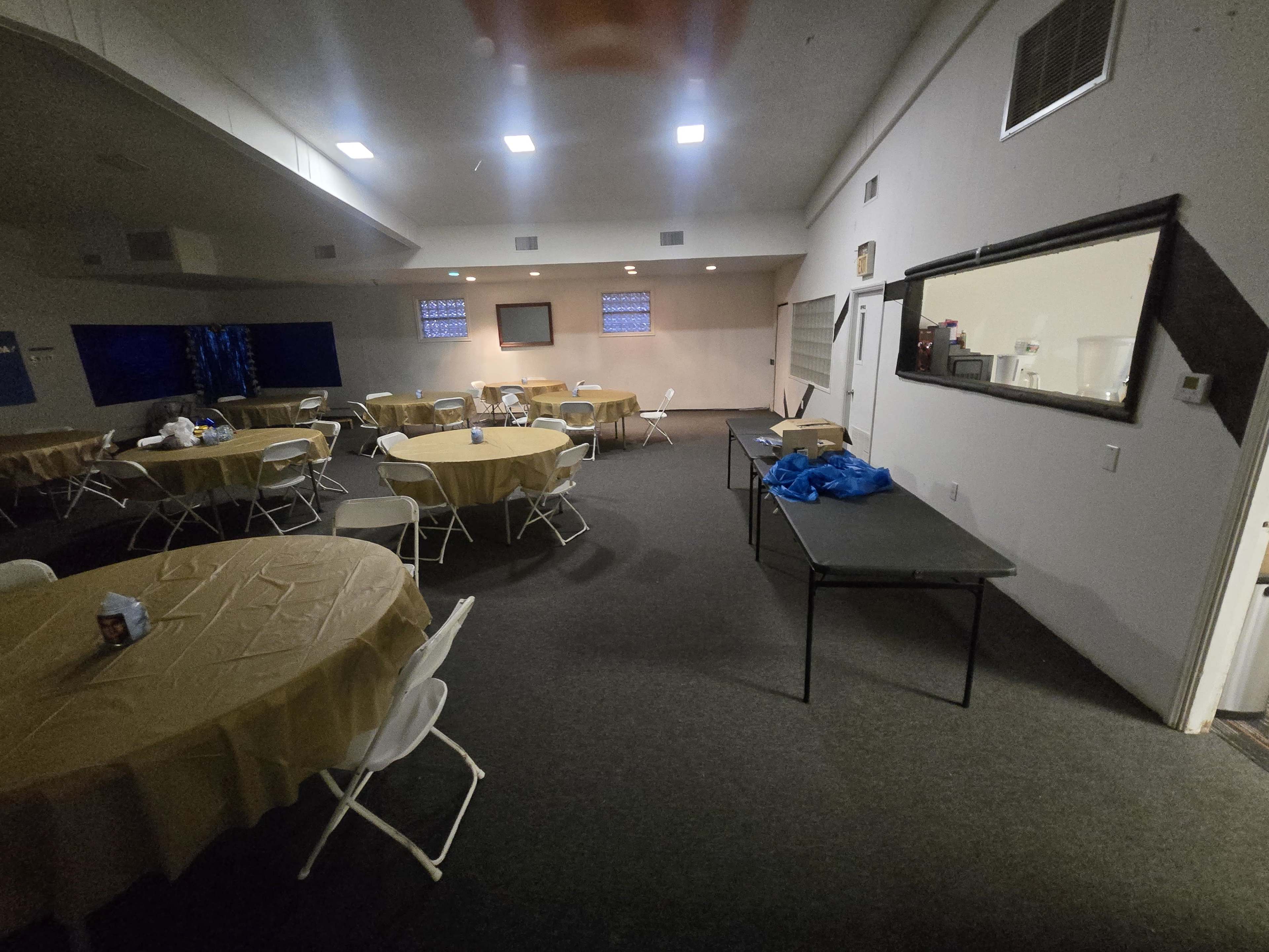 The image shows an empty room with several round tables covered in tan tablecloths and a long table along one wall, with some items placed on it.
