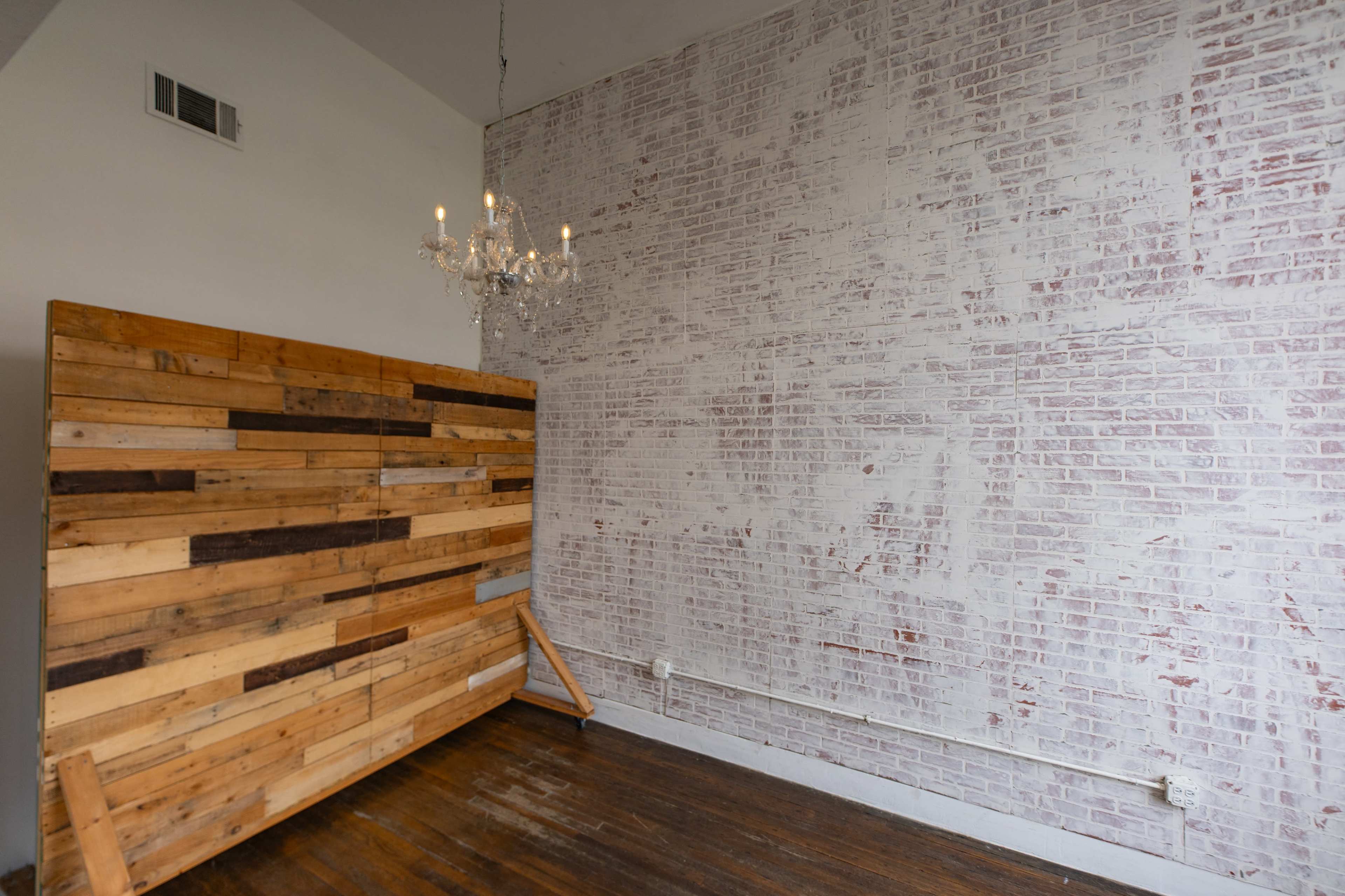 The image shows a room with a wooden accent wall on the left, a white brick wall on the right, and a chandelier hanging from the ceiling.