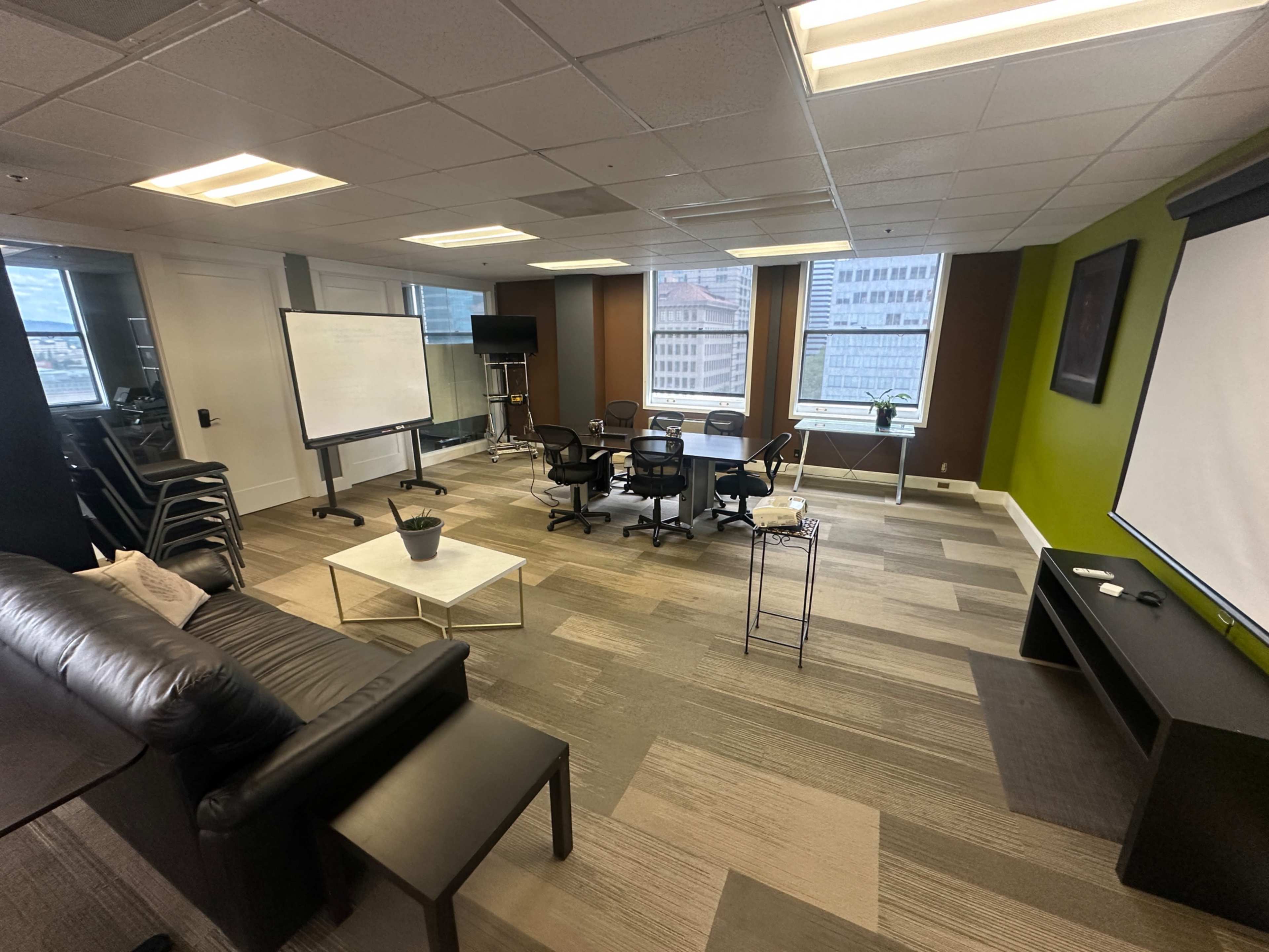 The image shows a modern office space featuring a seating area with a couch, a meeting setup with chairs and tables, and large windows providing natural light.