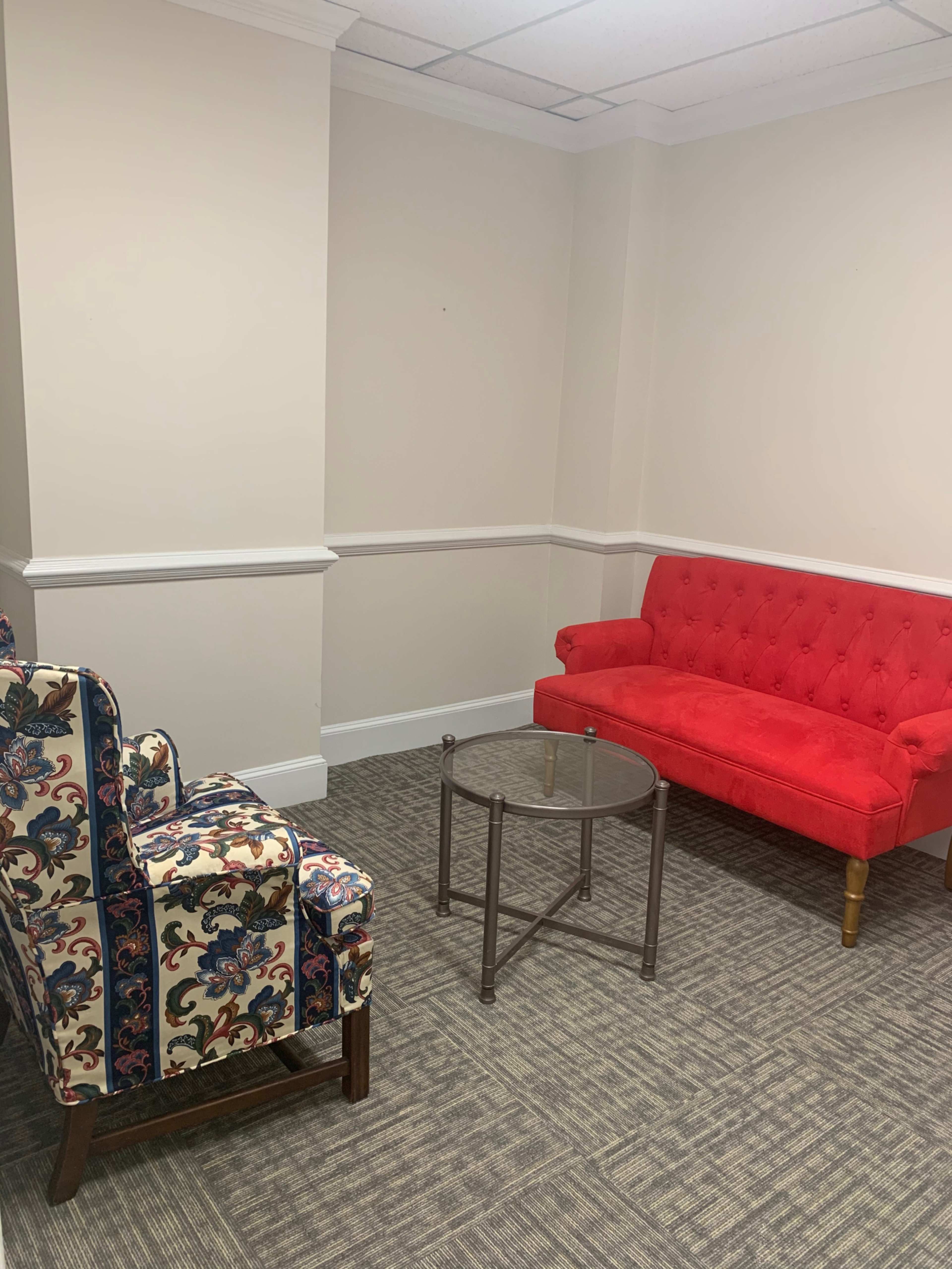 The image shows a corner seating area with a patterned armchair, a red tufted sofa, and a glass-top table on a carpeted floor.