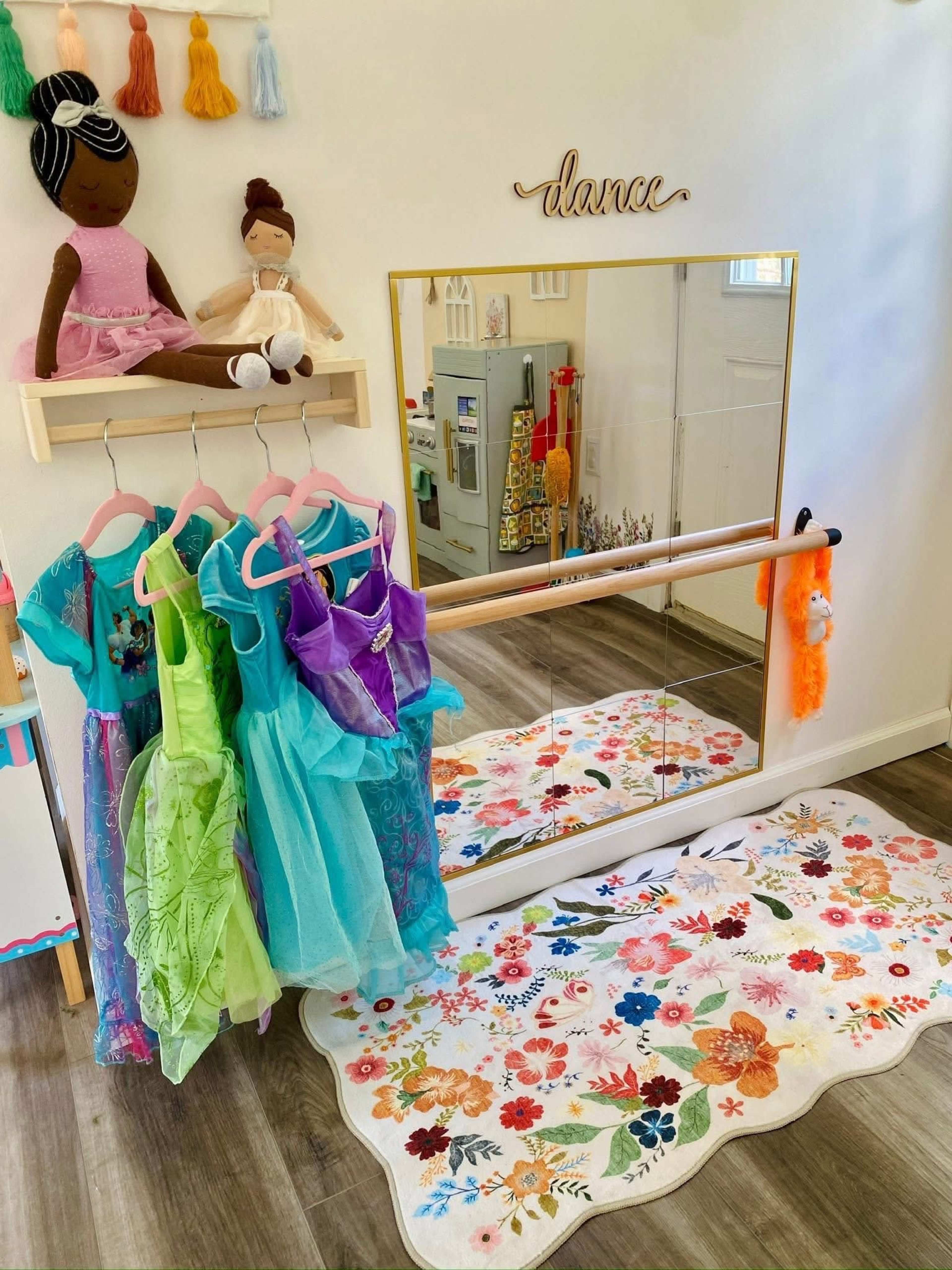 The image shows a children's dance area with colorful dresses hanging on a rack, a mirror, a shelf with a doll, and a decorative rug on the floor.