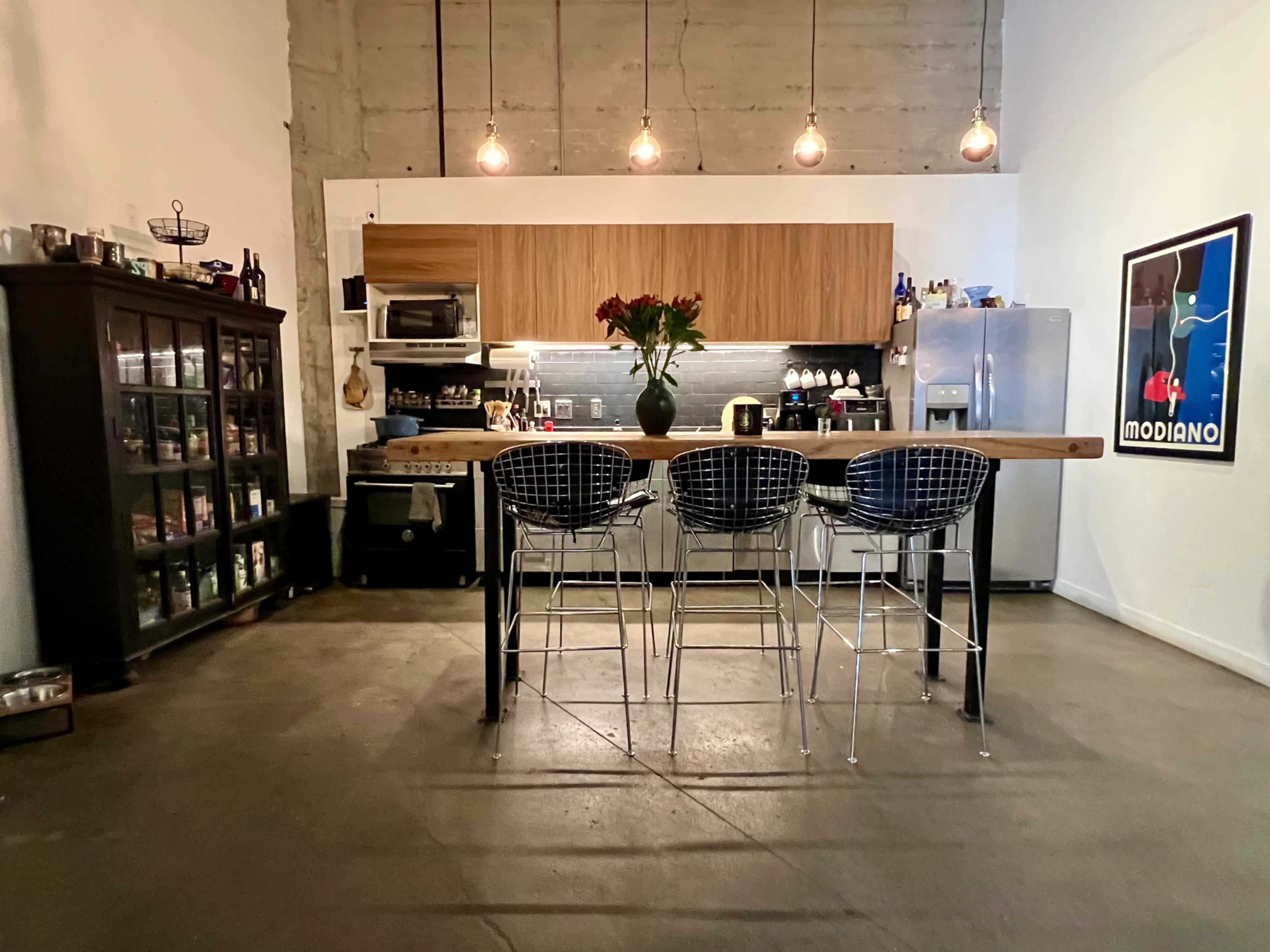 A minimalistic kitchen features a wooden bar with three stools, stainless steel appliances, a grey backsplash, and a large glass-front cabinet filled with jars.