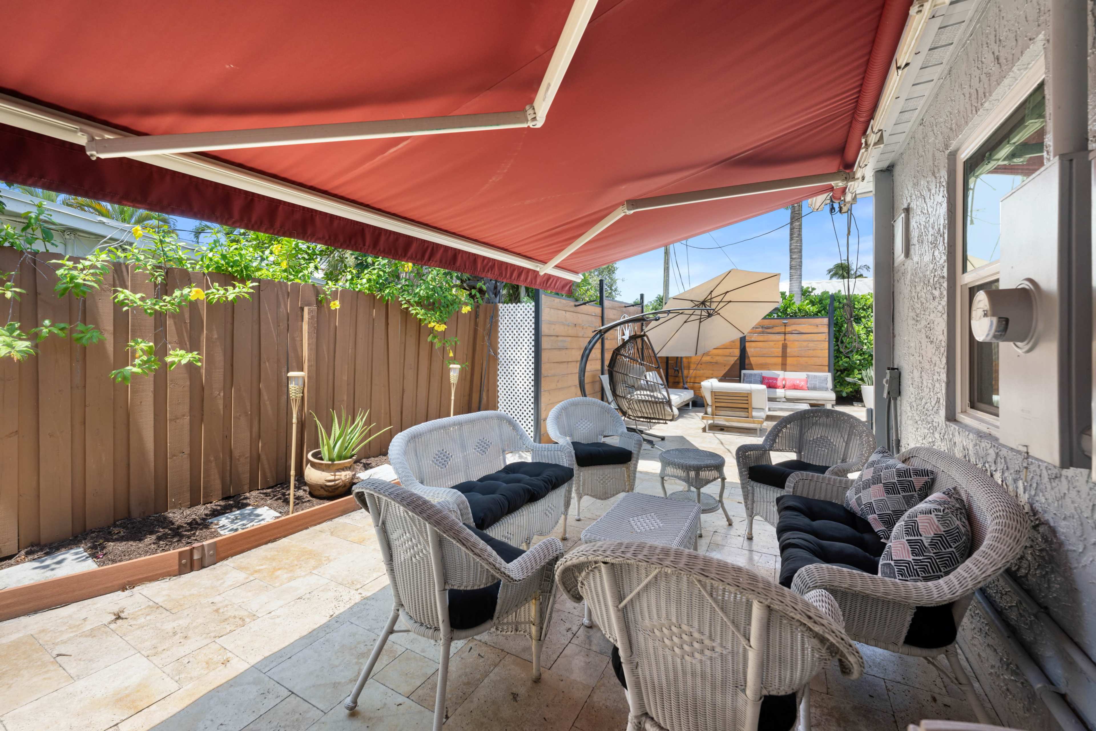 A covered patio features wicker chairs arranged around a circular table, with planters and a hanging chair visible in a secluded yard.