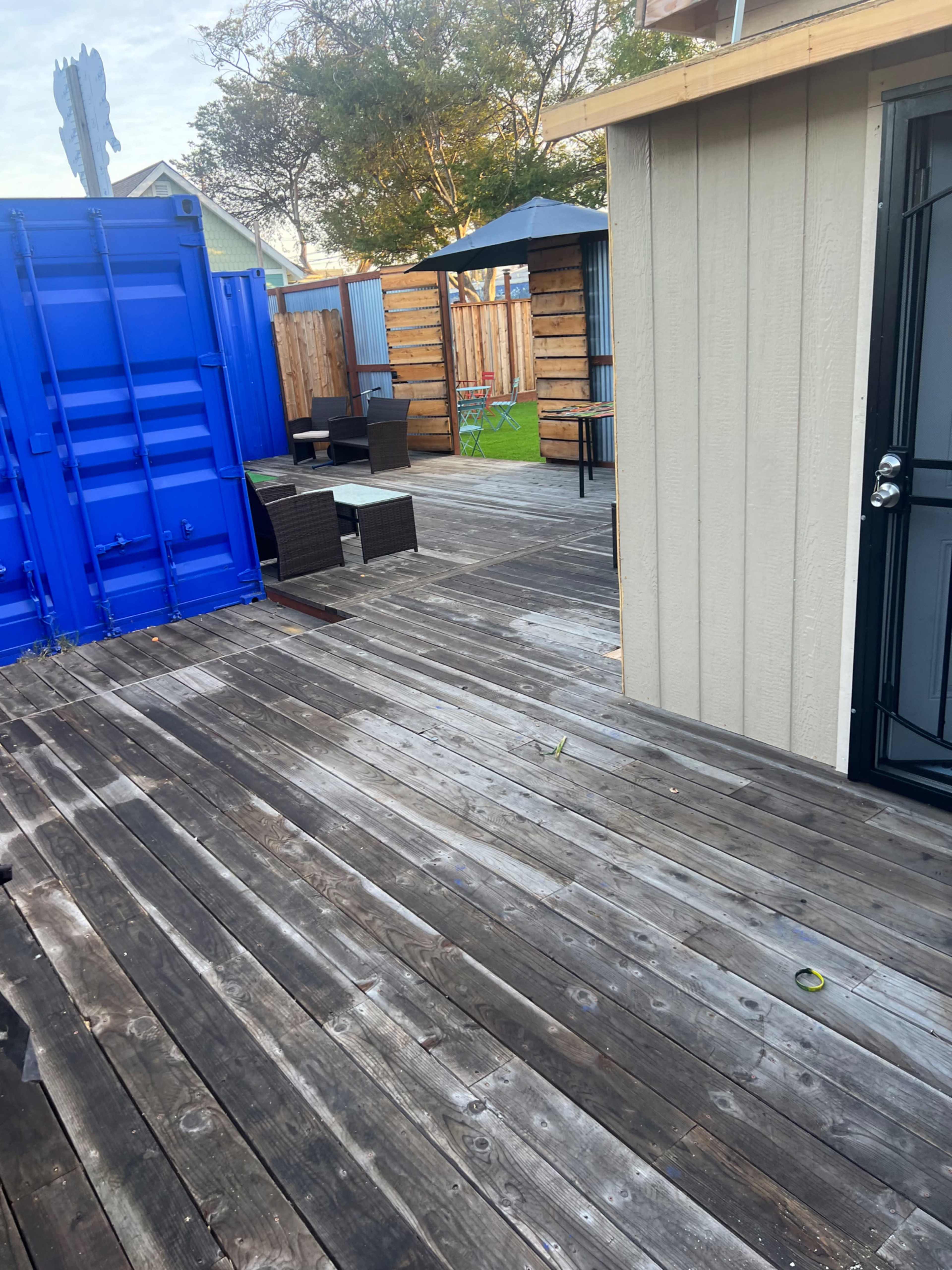The image shows a wooden deck area with seating furniture, a canopy, and a blue shipping container in the background.