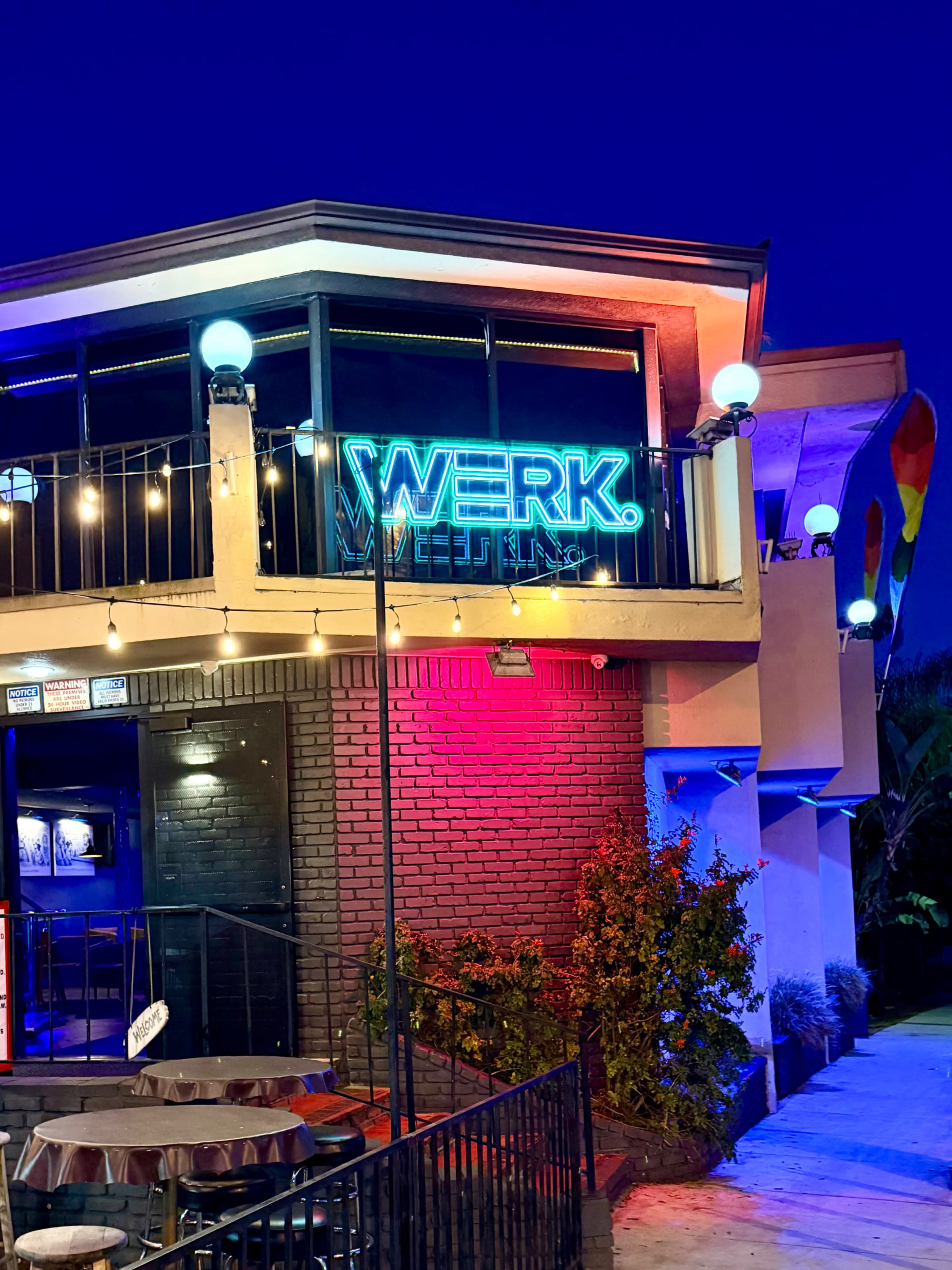 A brightly lit building displays a neon sign reading "WERK" surrounded by colorful outdoor lighting and a small patio area.