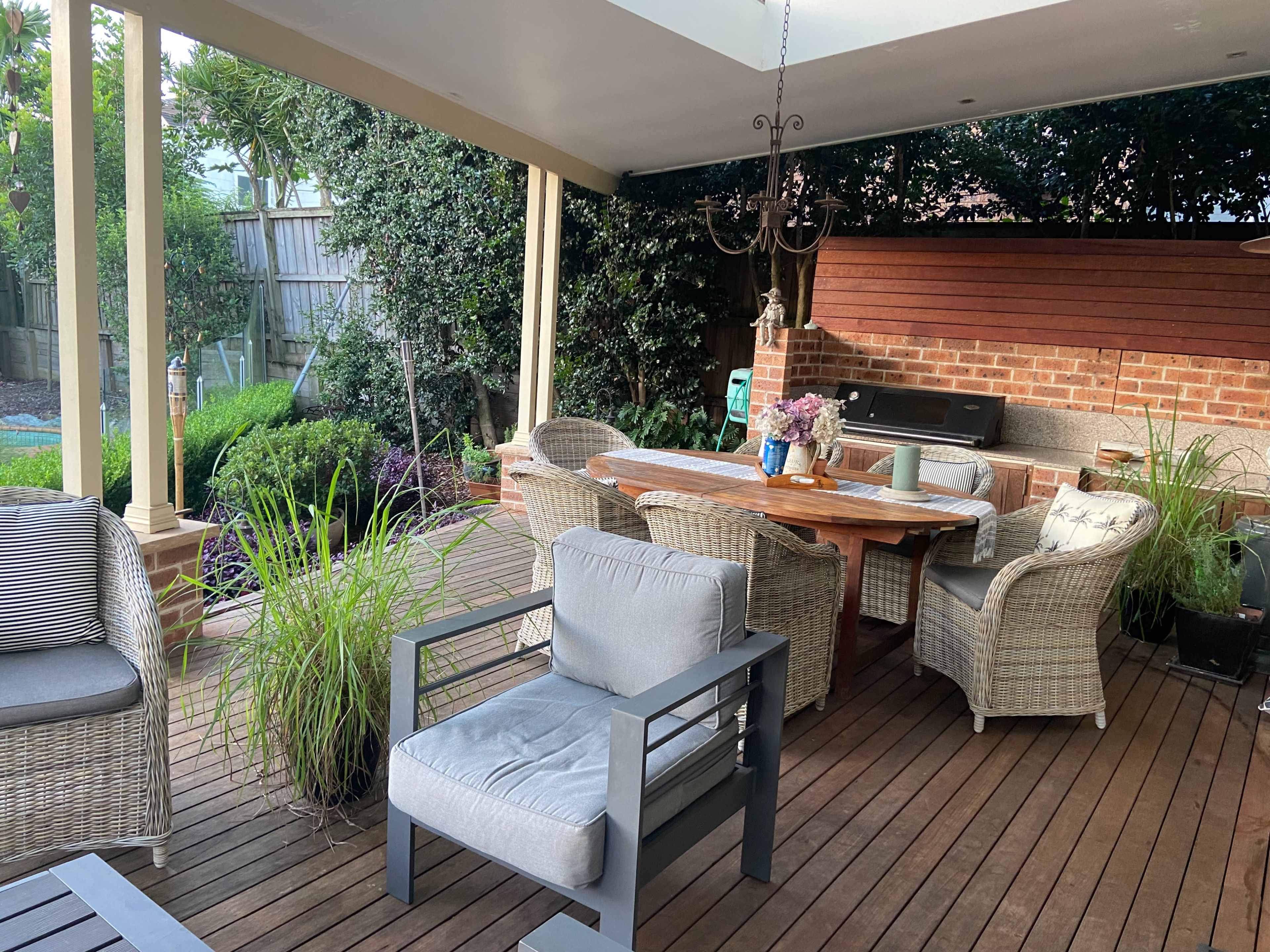 A spacious outdoor patio features a wooden dining table surrounded by wicker chairs, alongside potted plants and a barbecue grill under a covered roof.