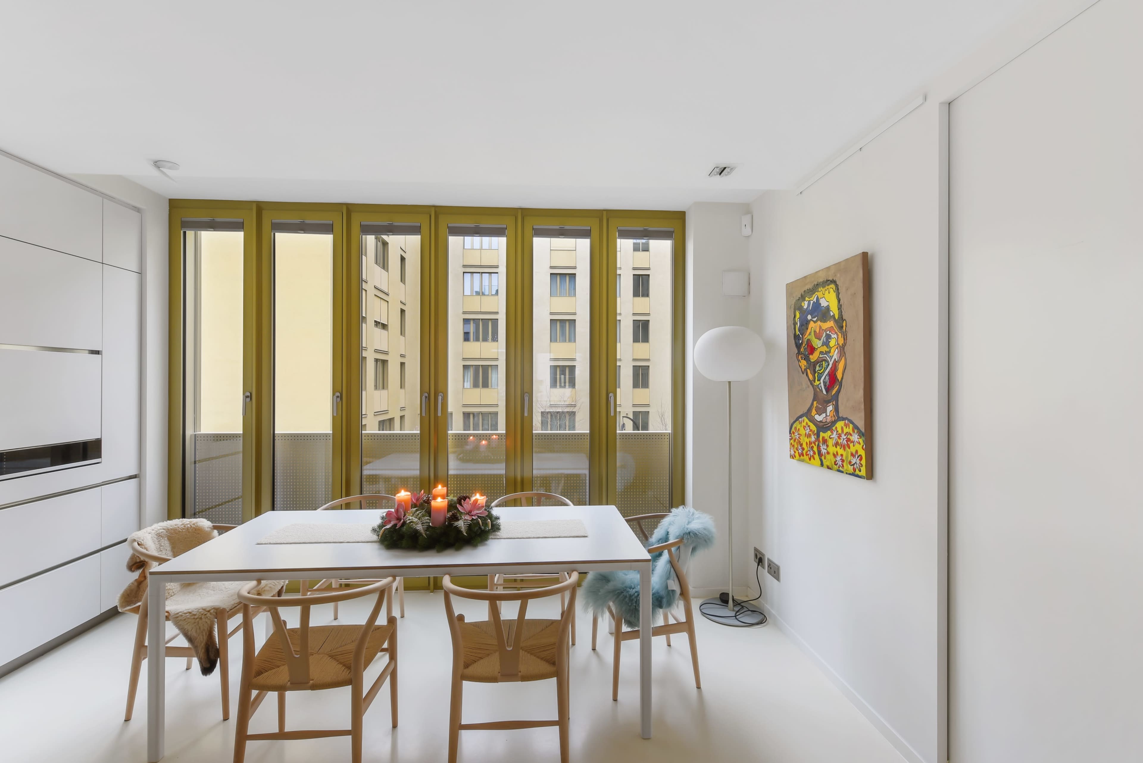 A minimalist dining area with a table set for four, featuring a centerpiece of candles, and large windows overlooking a building courtyard.