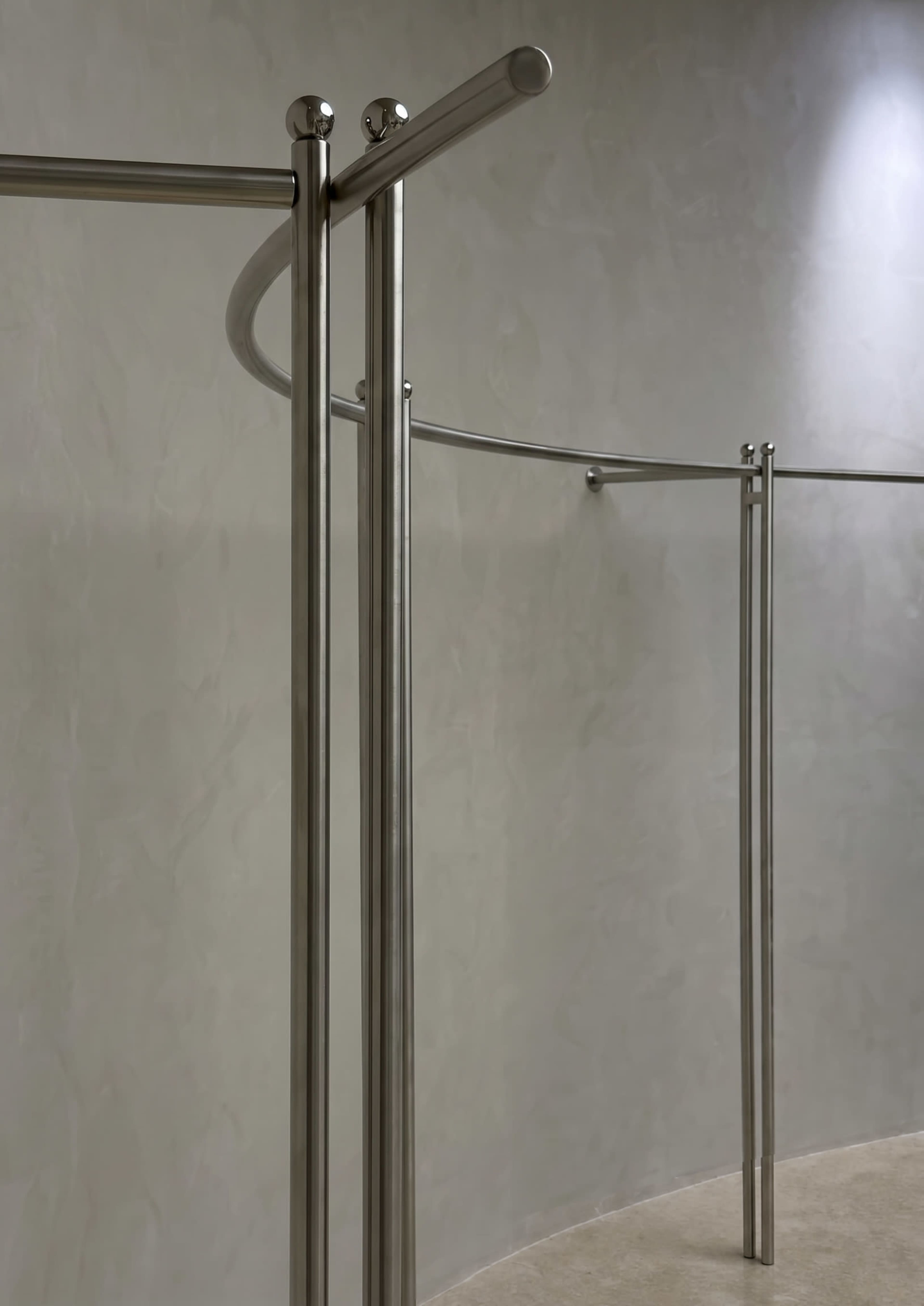 The image depicts a minimalist interior featuring curved stainless steel poles against a textured gray wall.