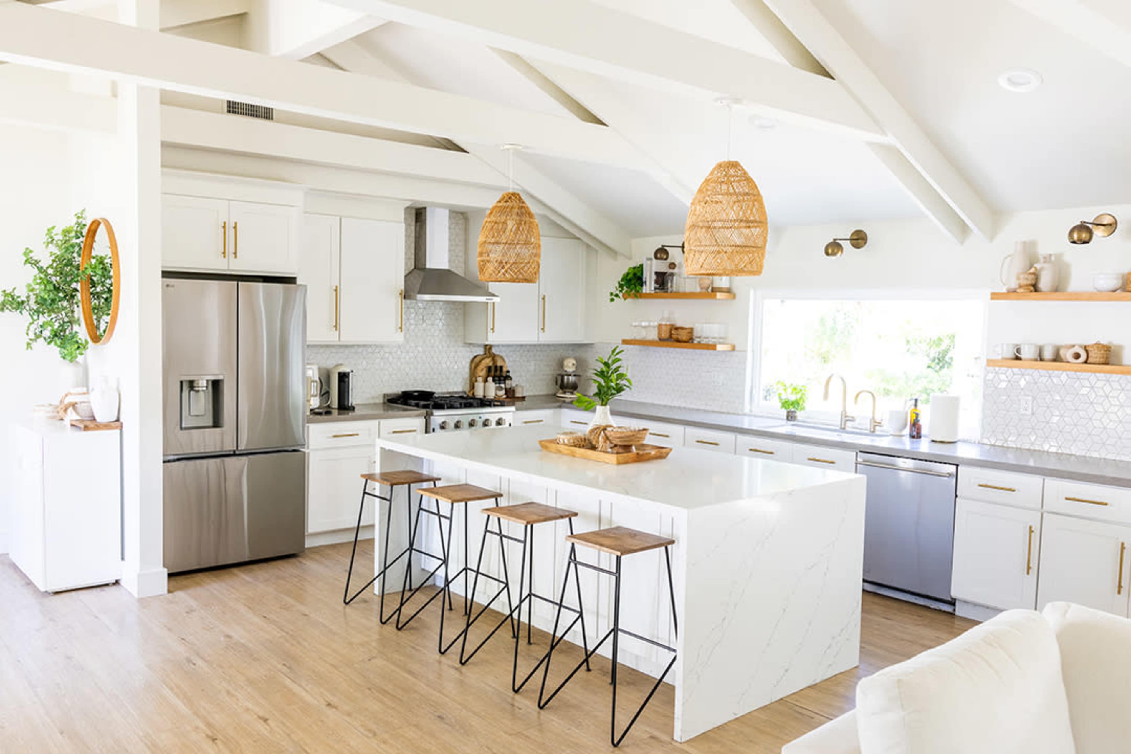 The image shows a modern kitchen with white cabinetry, a large island with barstools, and wooden accents, featuring stainless steel appliances and a bright workspace by a window.