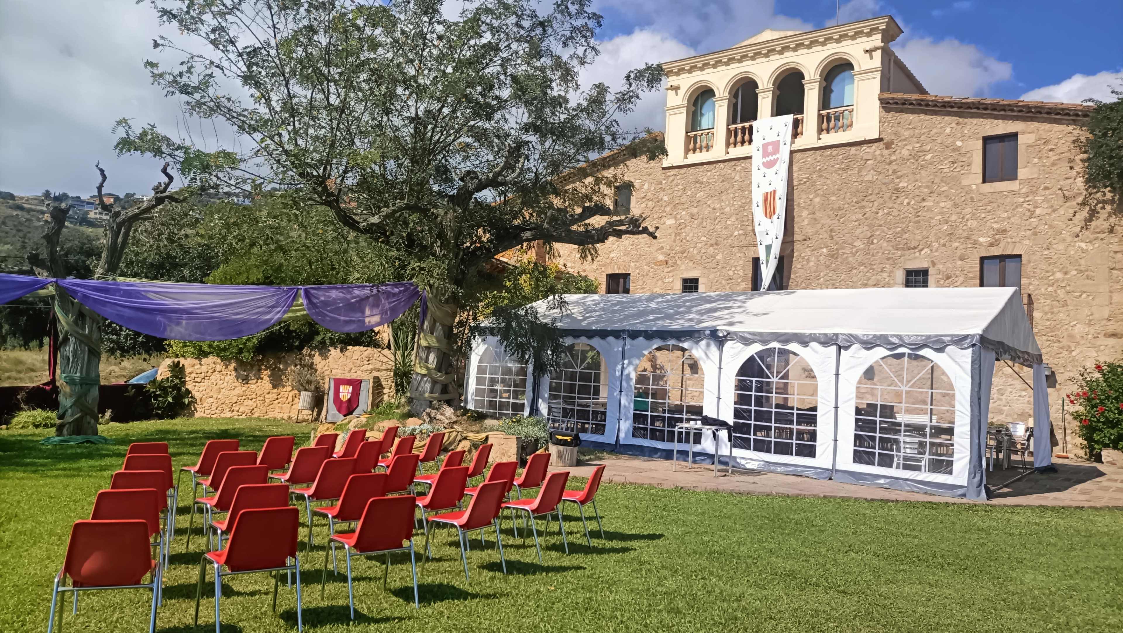 A wedding venue setup features rows of red chairs facing a large white tent beside a rustic stone building, surrounded by greenery.