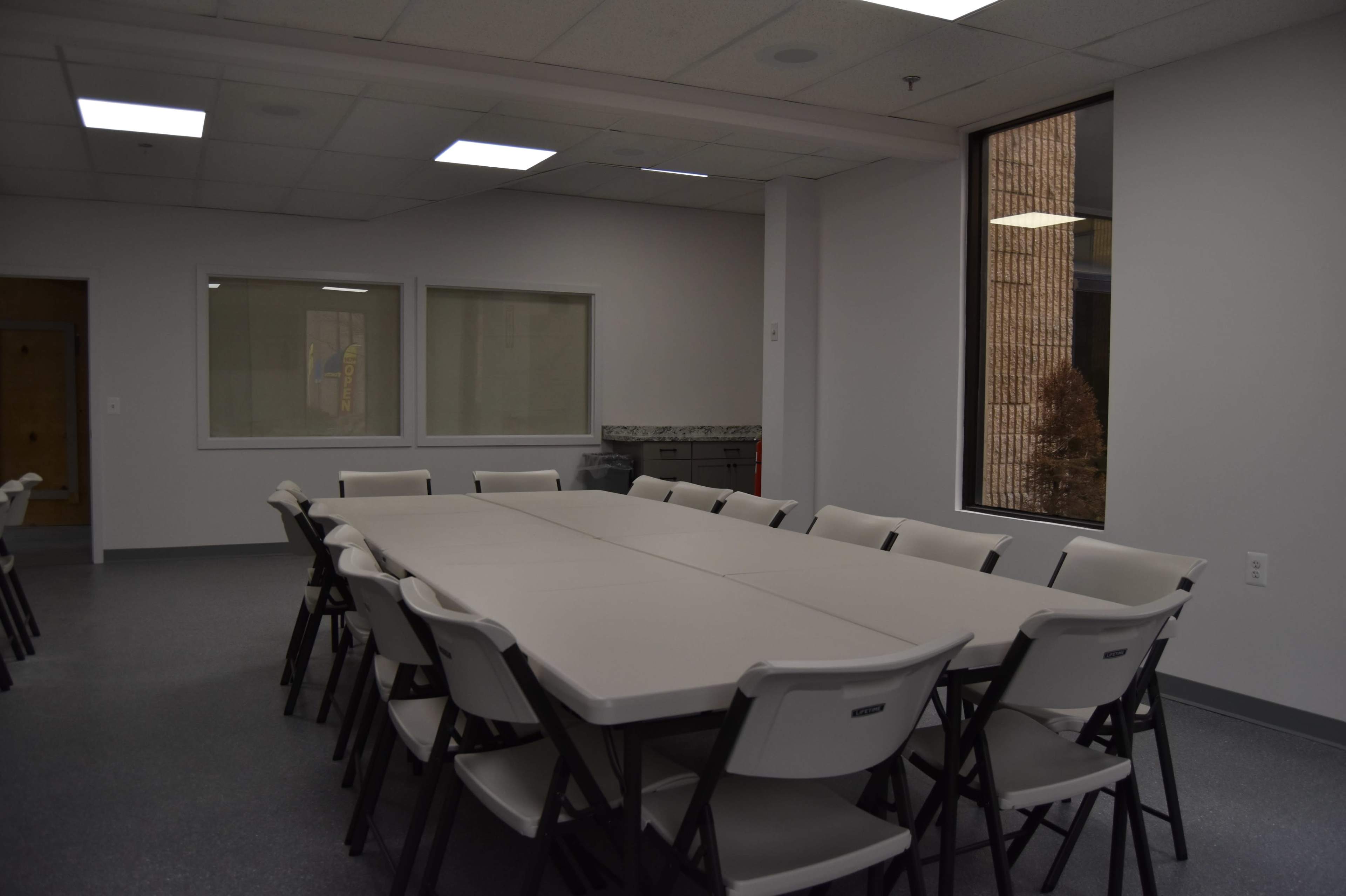 A meeting room with a large rectangular table surrounded by white folding chairs, featuring windows that provide natural light.