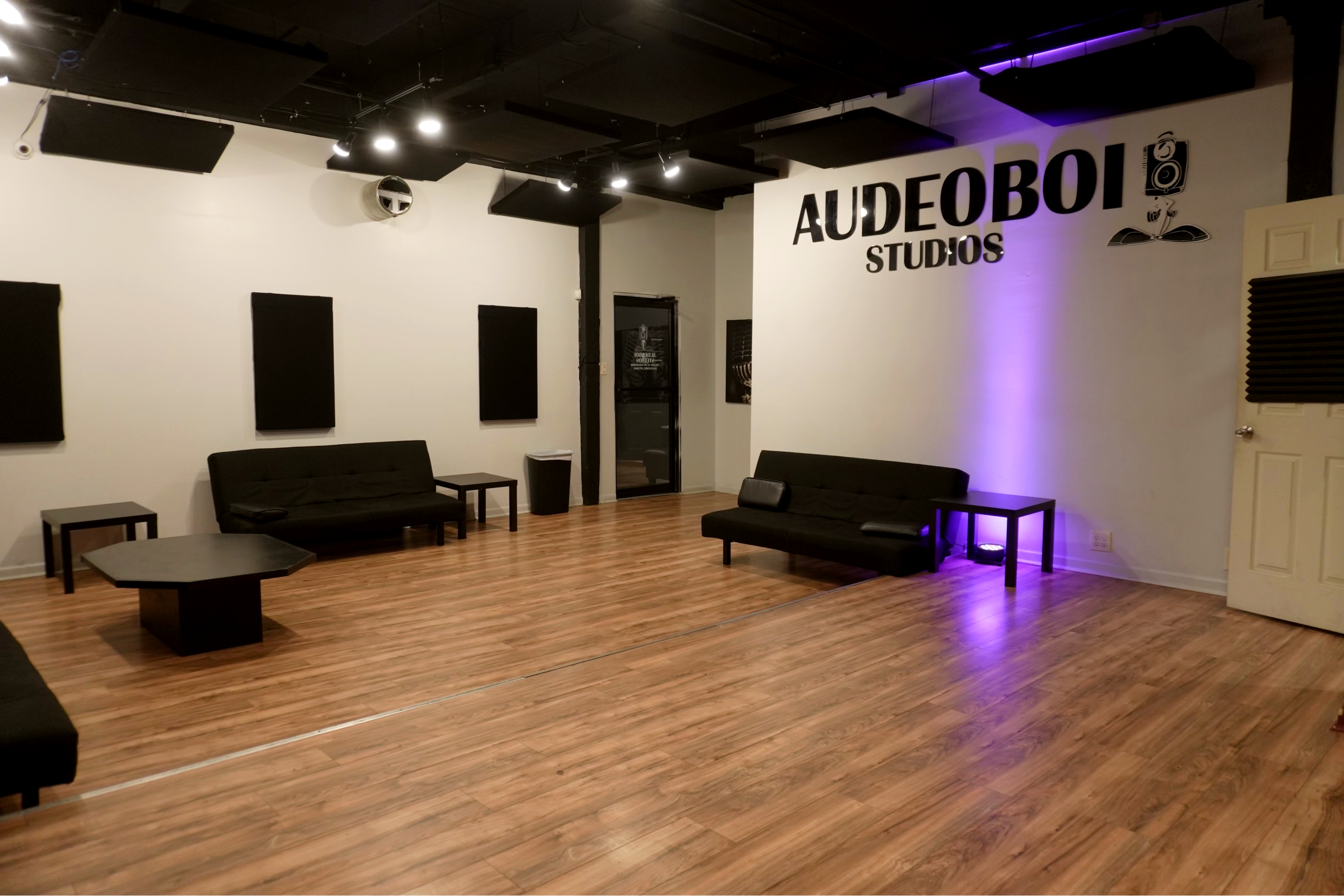 A modern studio space with black sofas, a central table, and soundproof panels on the walls, featuring the name "AUDEOBOI STUDIOS" prominently displayed.