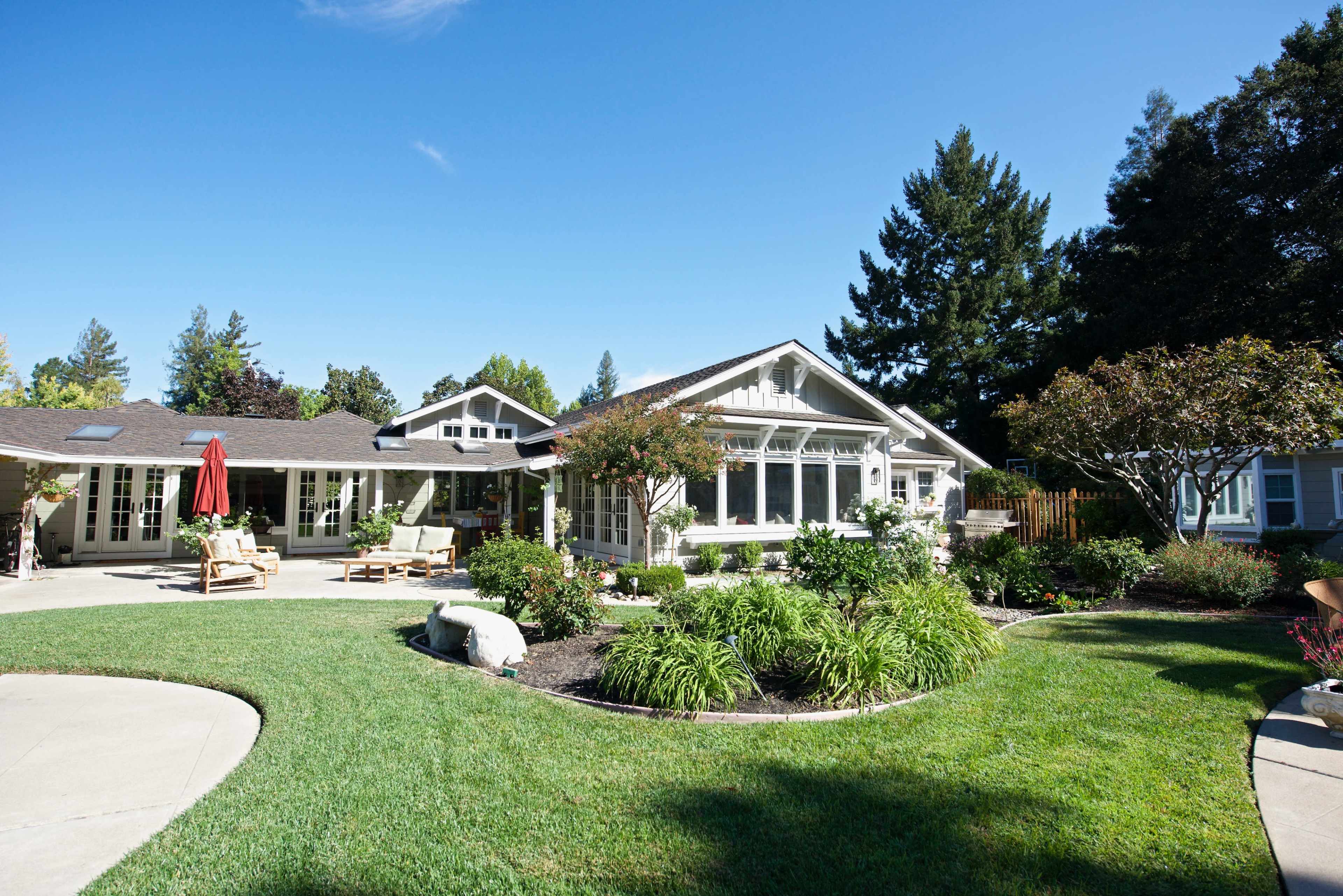 A well-maintained green lawn surrounds a large, modern single-story house with multiple windows and a covered patio area.